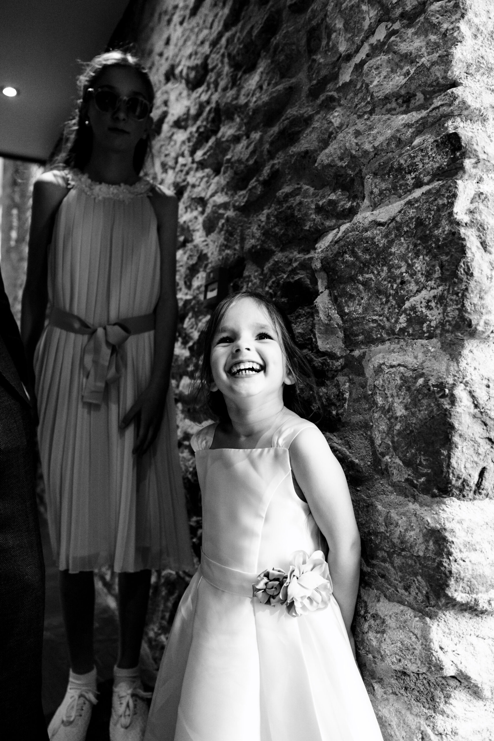 A black and white image of a little girl who is smiling showing all of her teeth and looking up to the camera. She is in a dress with a ribbon around the centre.