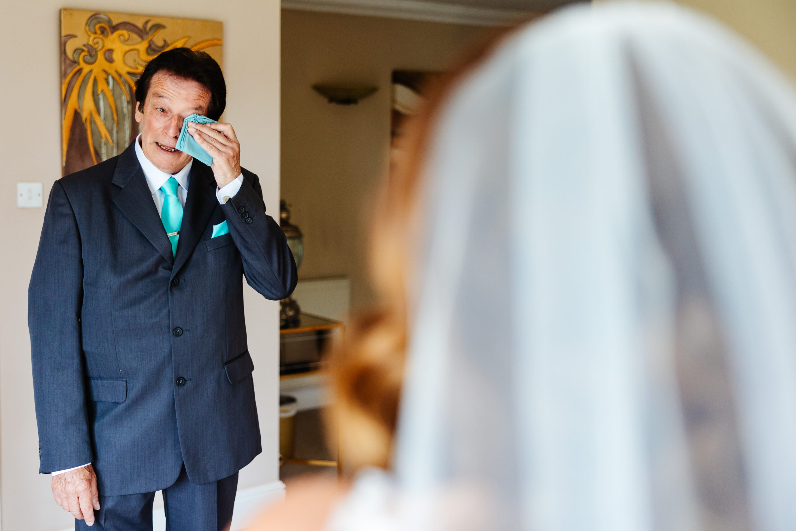 A man in a blue suit with a turquoise tie on. He is wiping a tear away from his eye.