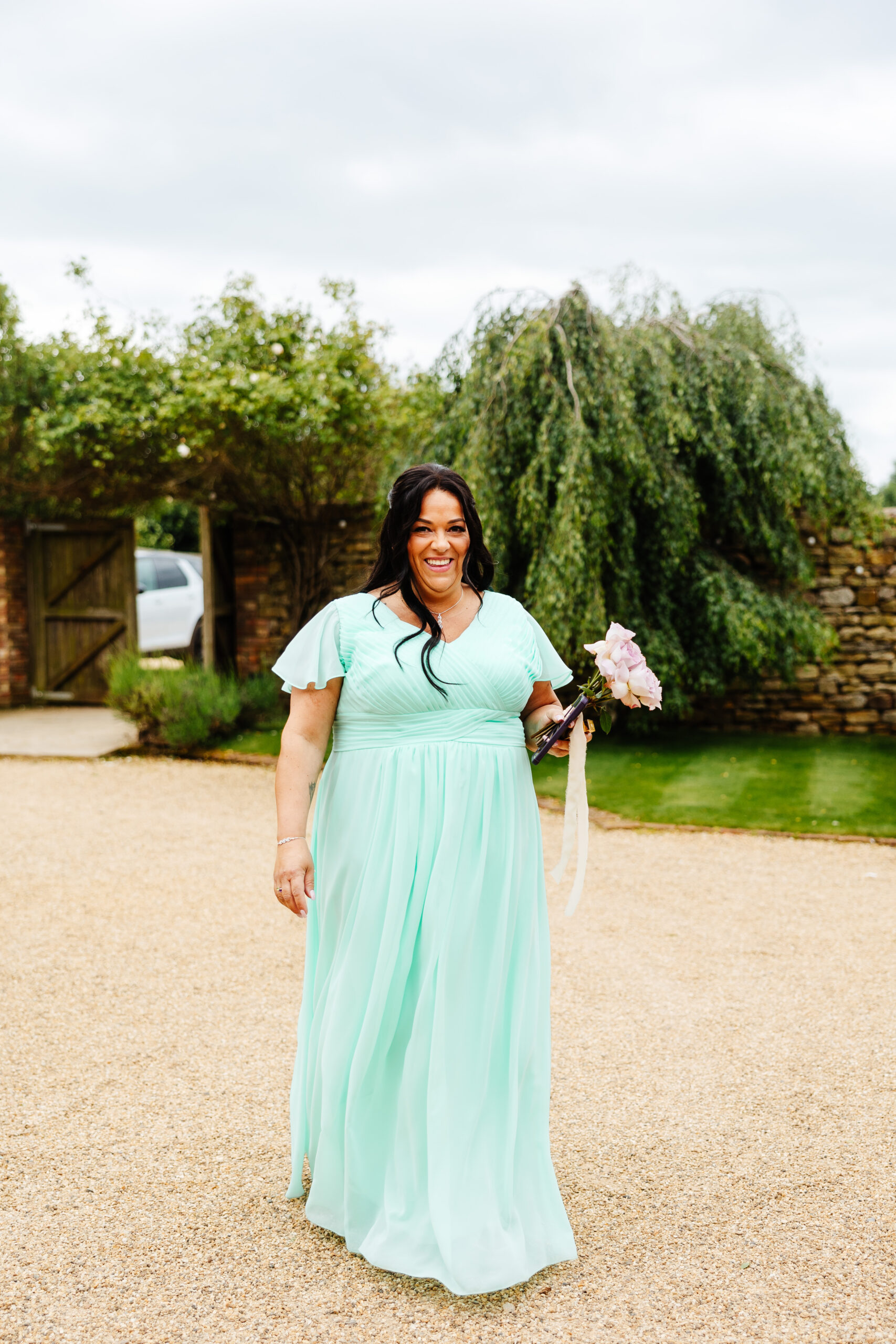 A photo of a bridesmaid in a turquoise dress. She is holding pink roses and her backdrop is a luscious green tree.