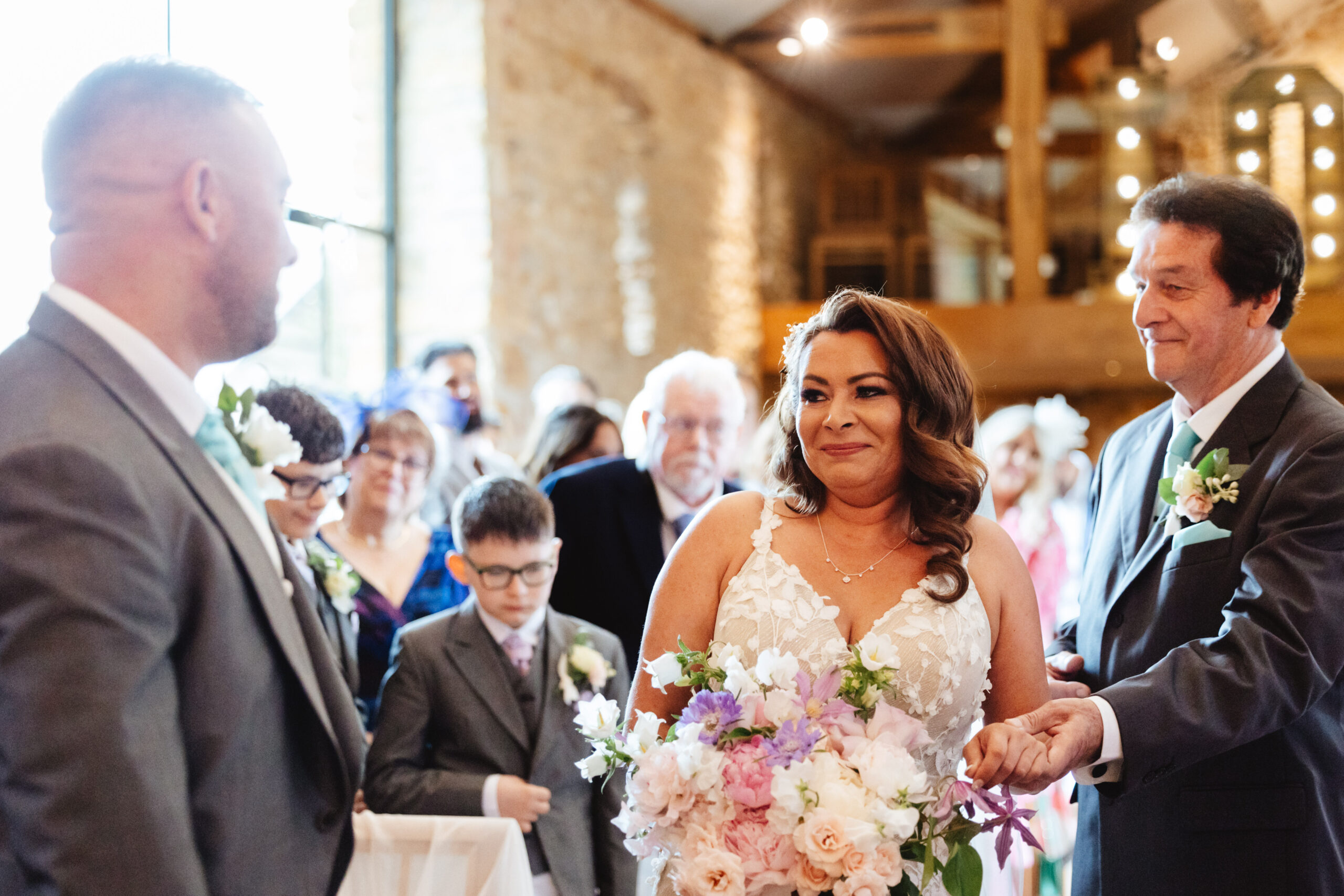 A photo of a bride and her father at the alter. Her Dad is giving her away to her groom. They are smiling and look emotional. The bride is holding a big bouquet of fresh flowers that are purple, white and pink. 