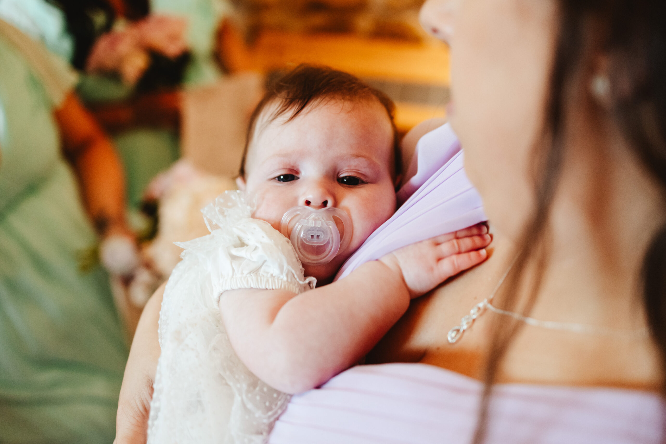 A photo of a baby in a white dress with a white dummy in. She is being held by a lady in a purple dress. The baby has her hand tucked in the dress on the lady's chest.