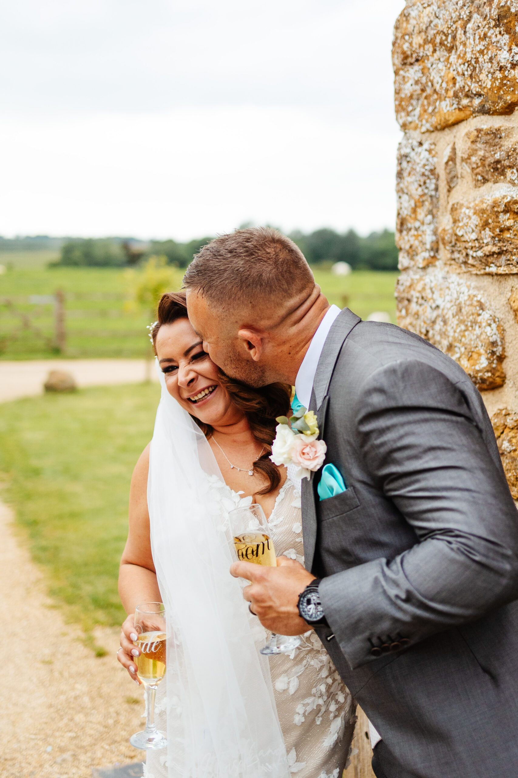 A bride and groom outside. The groom is giving the bride a kiss on her cheek and she is smiling with her nose crinkled. They both have a glass of champagne in their hand.