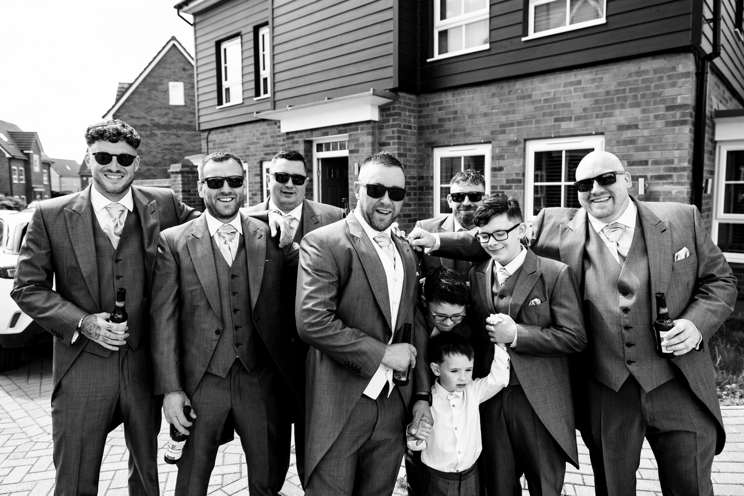 A black and white image of six men and three boys in grey suits and white shirts and are huddled in smiling at the camera.