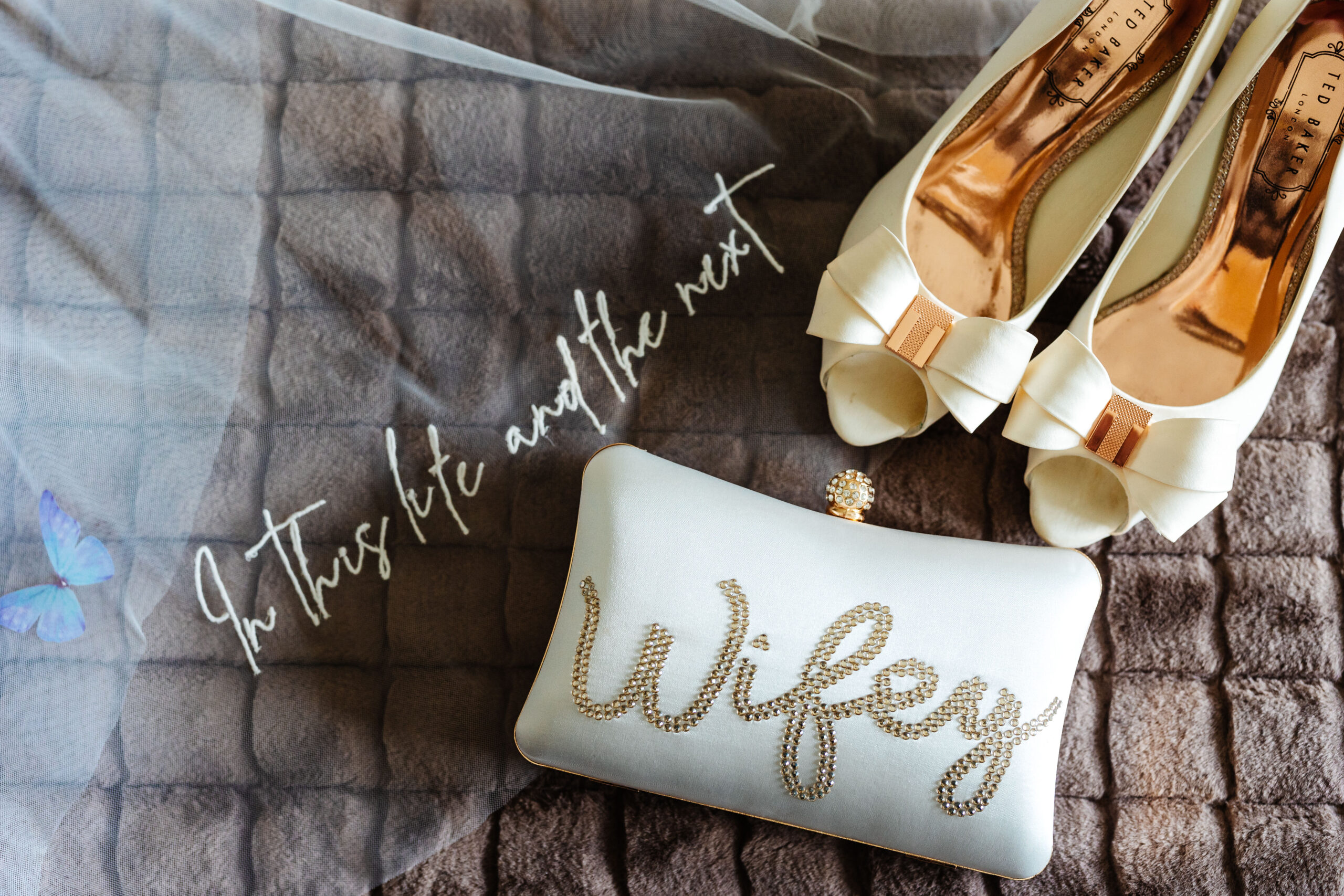 A photo of a veil with white embroidery on it saying "In this life and the next", a white pair of open toed shoes with a bronze bow in the middle and a white clutch bag with Wifey jewelled on it.