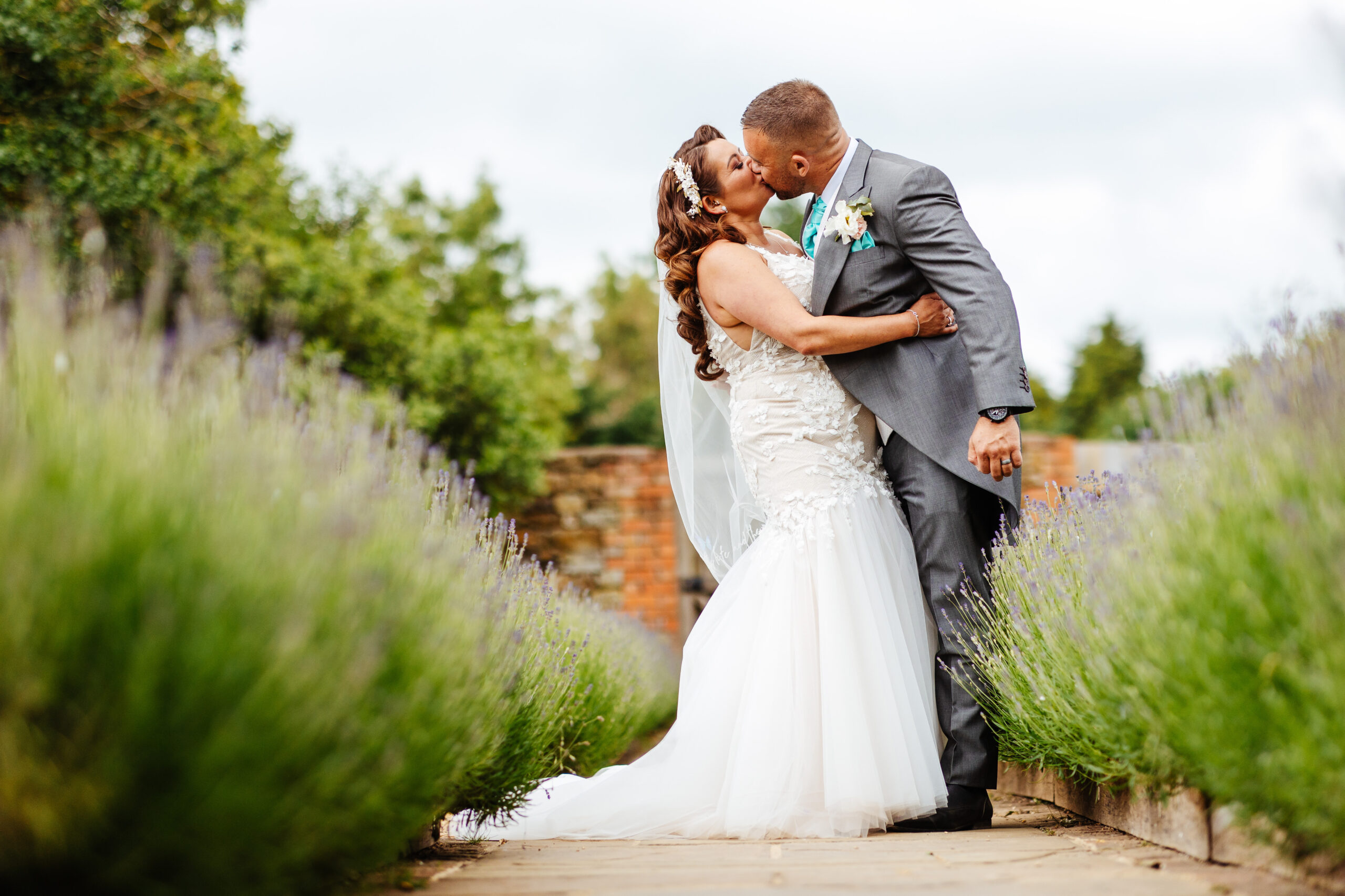 A photo of a bride and groom outside in a garden. The bride has a puffy white dress on, brunette hair flowing down her back and a veil on. The groom has a grey suit on with a turquoise tie on. They are kissing and the bride has her arm around his waist.