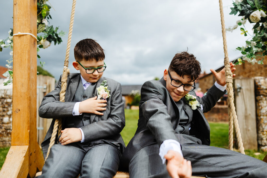 Two boys sat on a wooden swing. They have grey suits on and look like they are having fun.