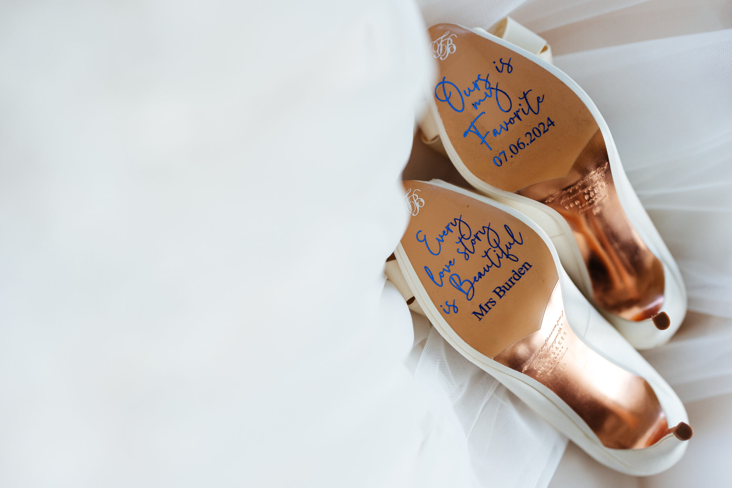 A pair of white shoes next to a white dress and the bottom of her shoes have blue printing on them with writing on. 
On the right shoe is Ours is my favourite and on the left shoe is Every love story is beautiful and Mrs Burden written on it.