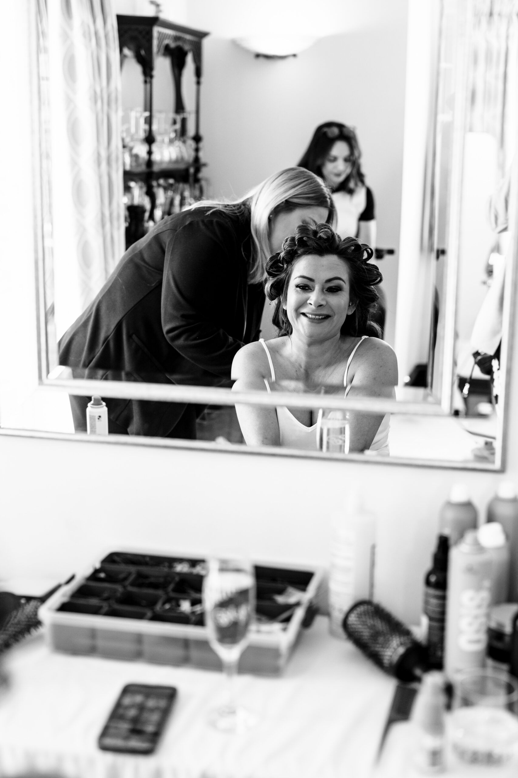 A black and white photo of a bride looking in the mirror. She has rollers in her hair and a lady helping her. She is looking at herself and smiling.