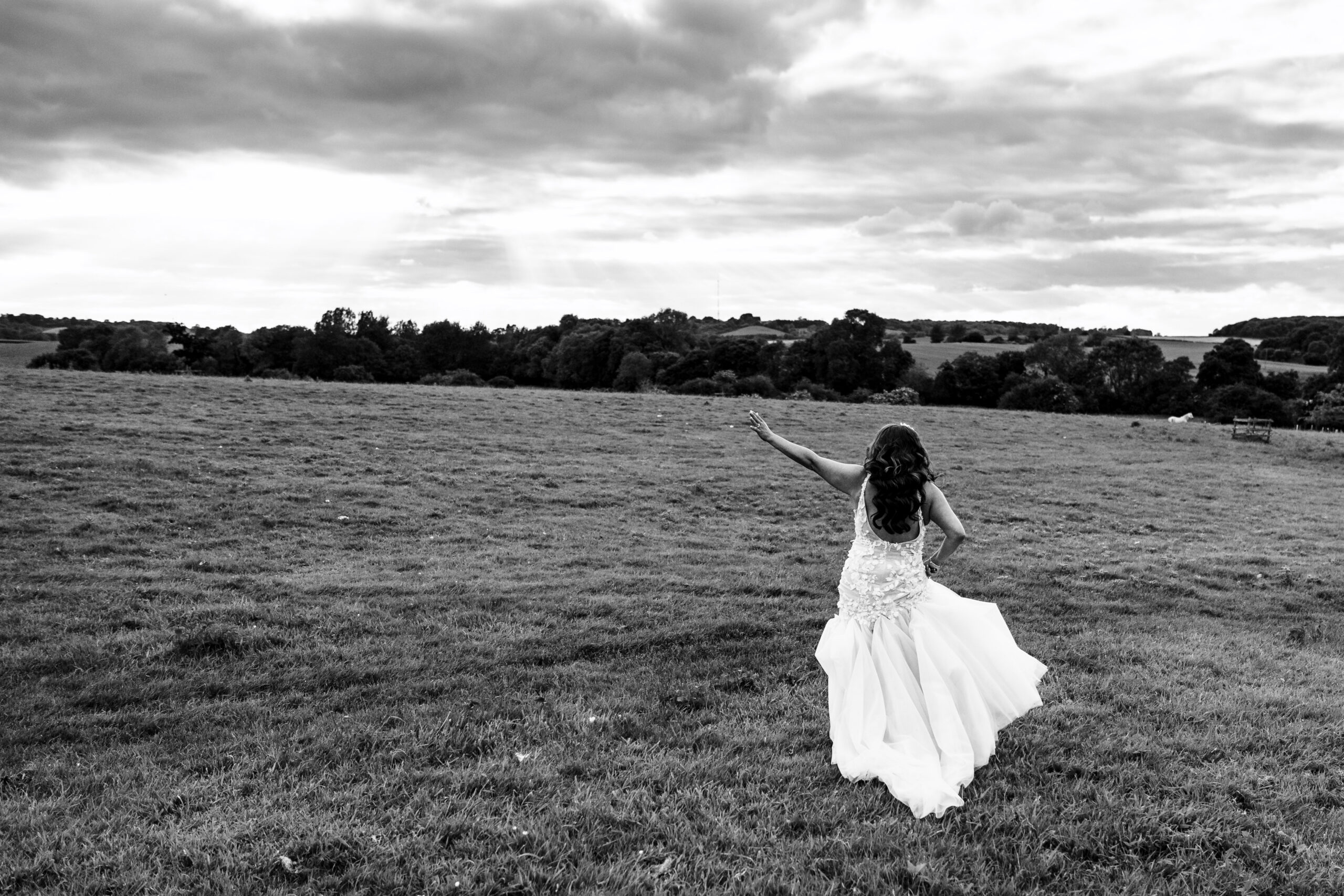 A black and white photo of a bride in a field on her own. She has her left arm in the air and her right hand is hitching her dress up