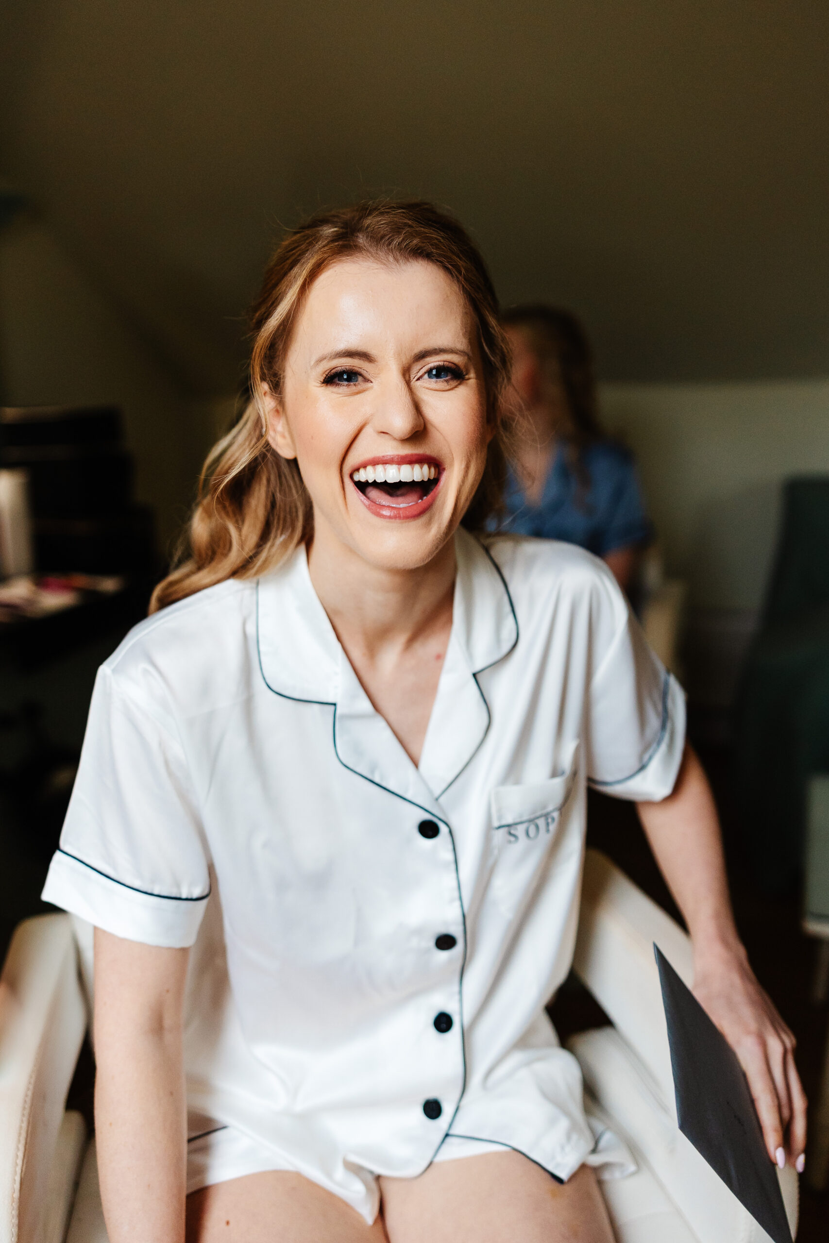 A photo of a bride in white pajamas with black trim and black buttons. Her blonde hair is tucked behind her face, she has wedding make up on and she is smiling looking at the camera.