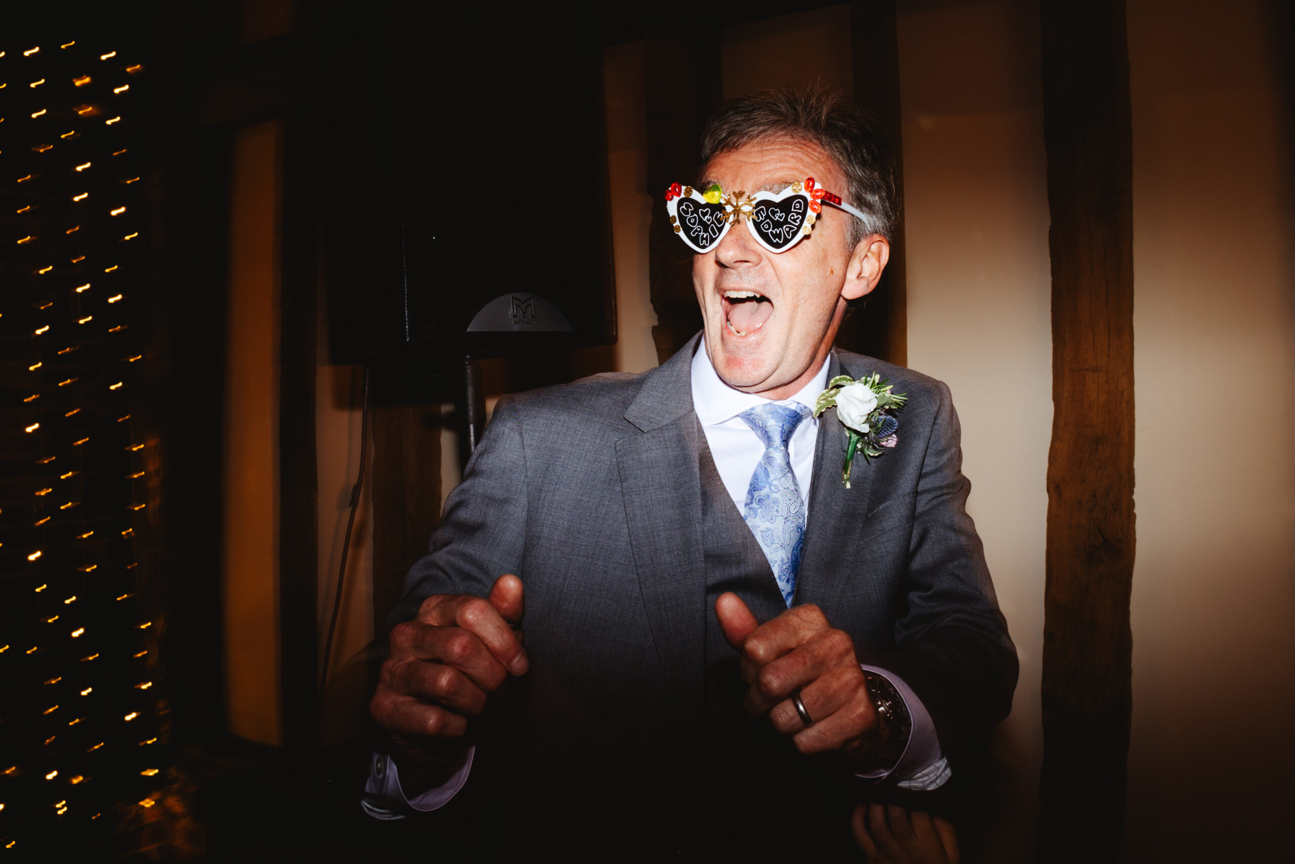 A man in a grey suit with blue detailing (tie) wearing white, heart sunglasses that are decorated with bright coloured stickers on and Sophie and Edward written on the lenses. He has a happy face and is dancing.