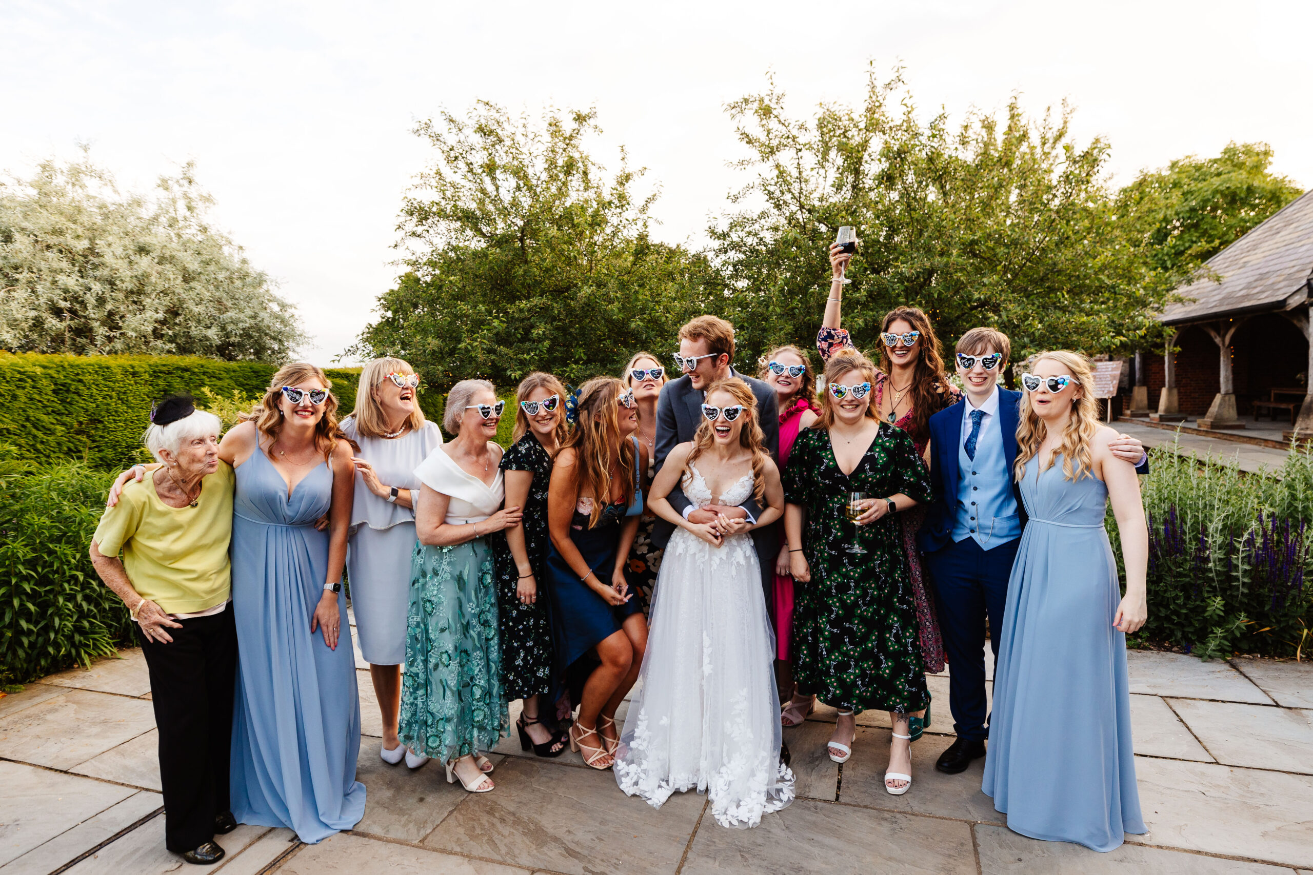 A group image of family and friends surrounding the bride and groom outside. They all have decorated, heart sunglasses on and are having a good time.