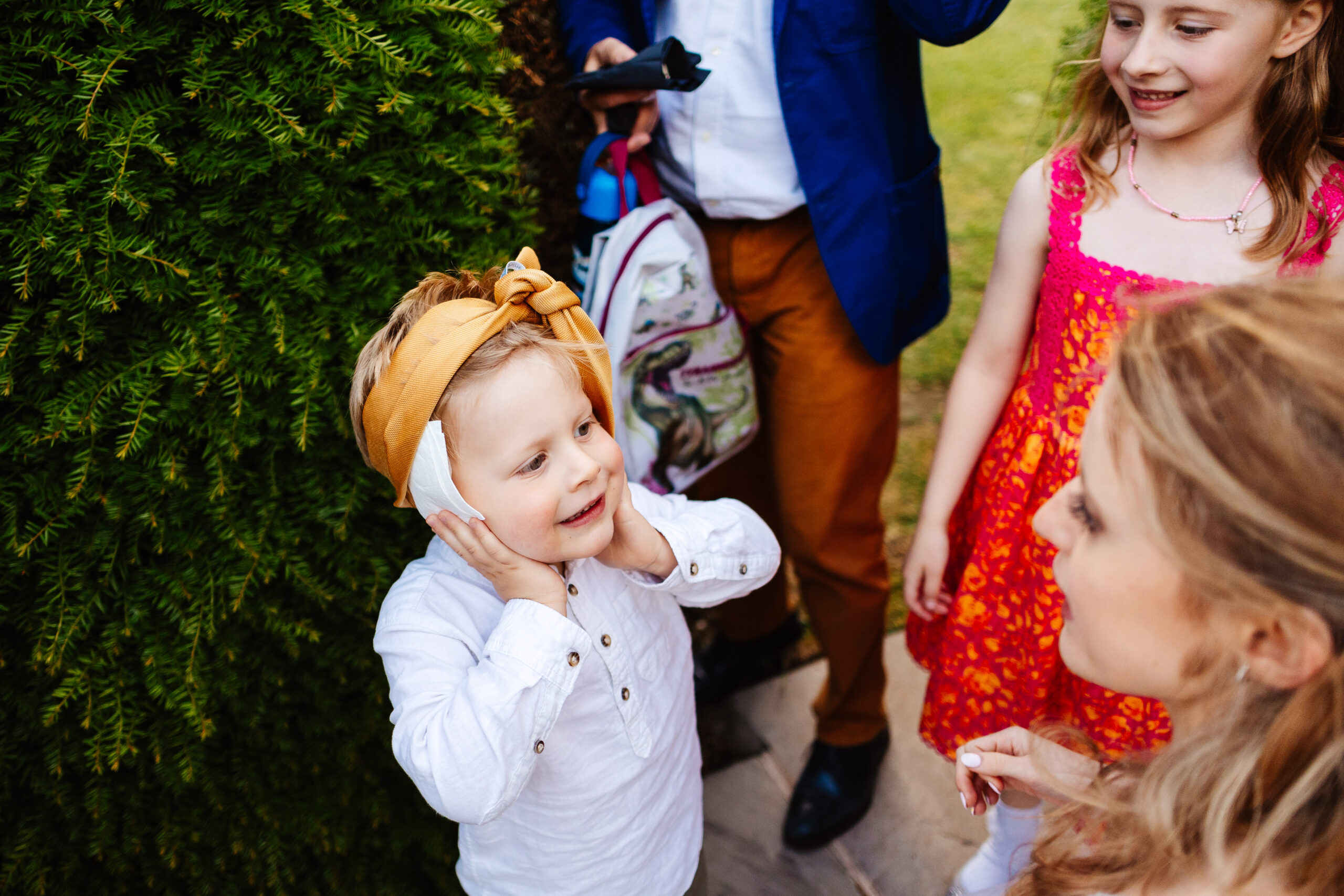 A little boy in a white shirt with a headband around his head. He is with the bride and another little girl who are looking at him and smiling.