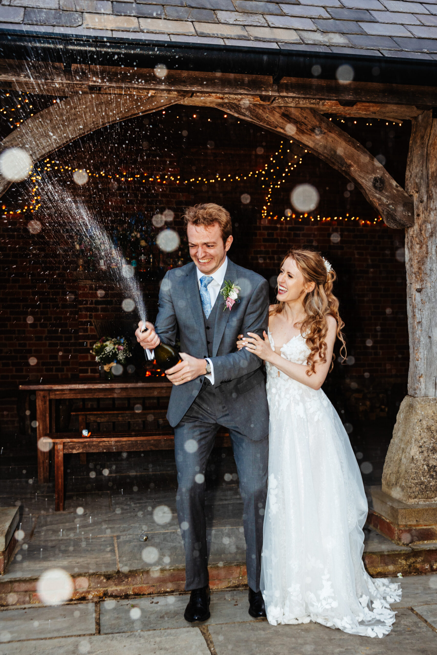 A bride and groom outside near a bar spraying a bottle of champagne. You can see the champagne shower all around and they are smiling and laughing.