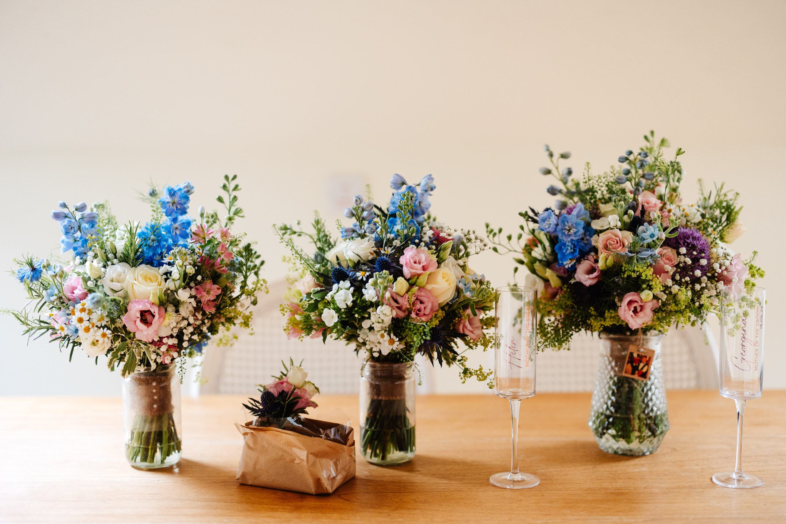 Three bouquets of flowers in clear glass jars. They are full of pastel pink and white flowers with big bursts of bright blue. There are also two champagne flutes on the table where the flowers are. They are engraved with people's names on.