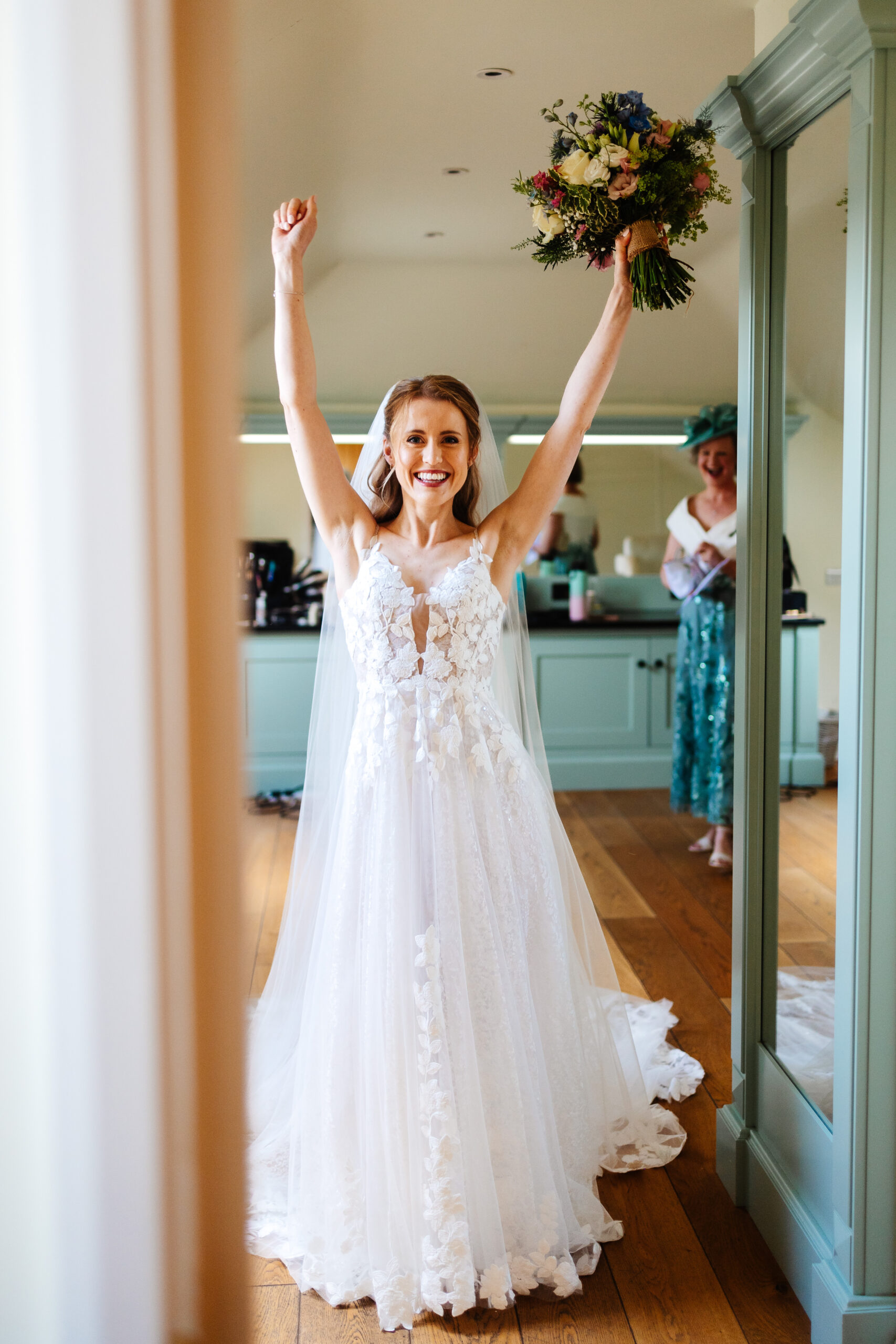 A photo of a bride in a detailed, lace wedding dress and veil. She has both arms in the air in excitement and in one of the hands she is holding her bouquet. She has a very excited face!