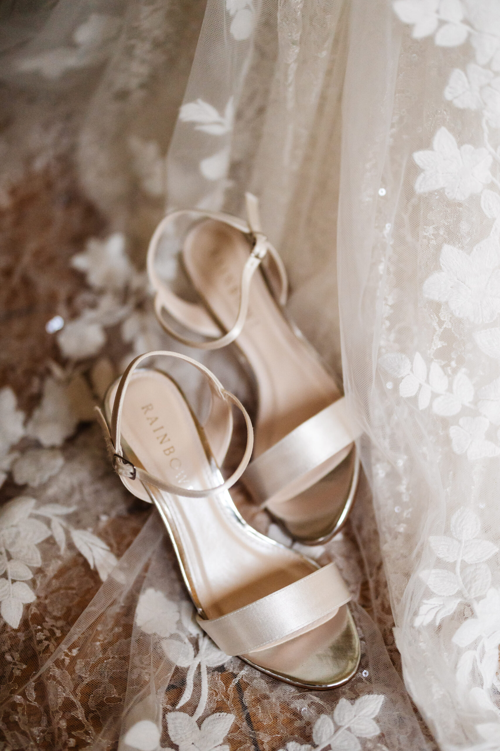 A pair of open toed, cream shoes on top of a laced bottom of a wedding dress.