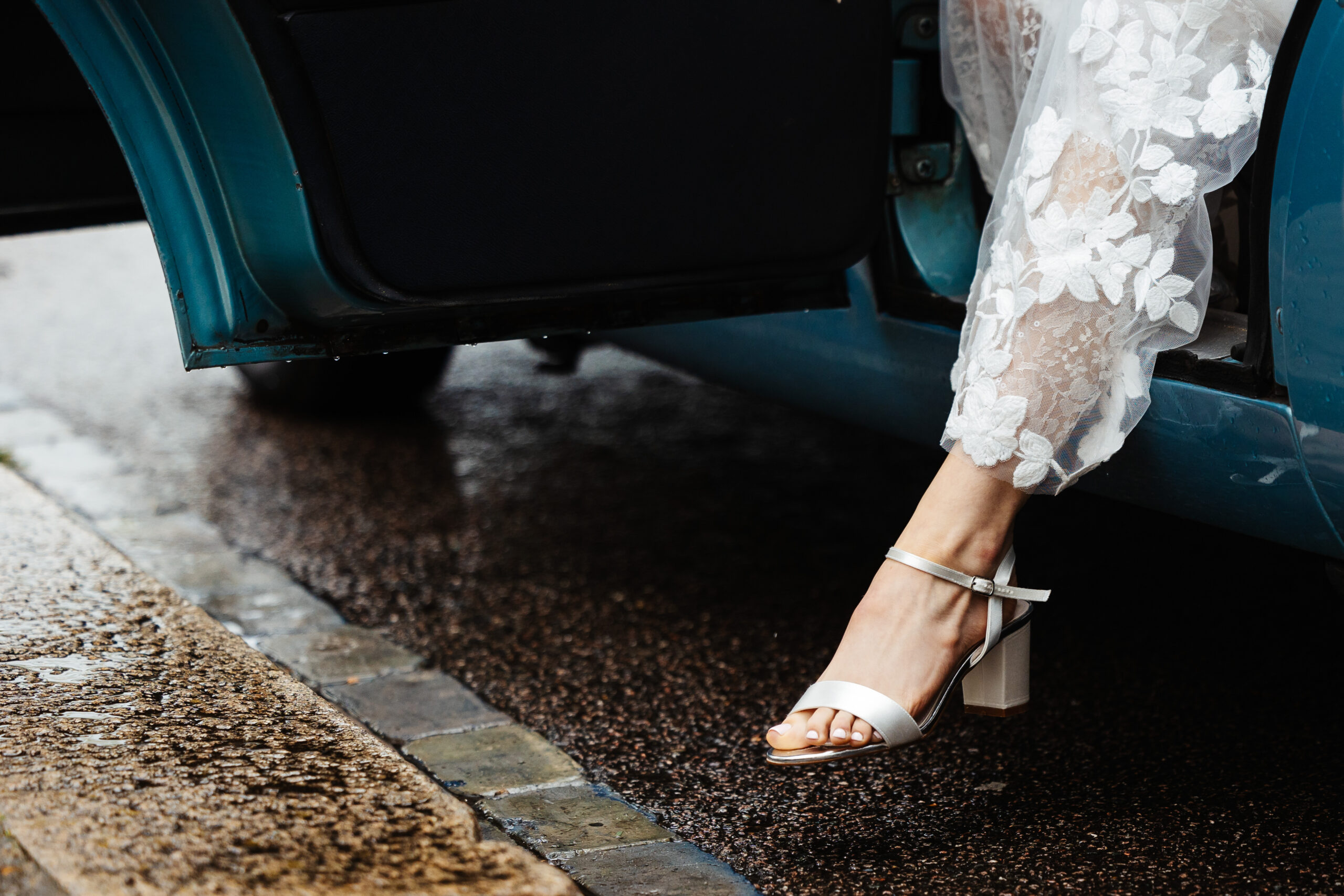 A blue car door is open and you can only see the foot and the leg of the bride. You can see her foot in an open toed cream heeled shoe and the bottom of her lace, detailed, white dress.