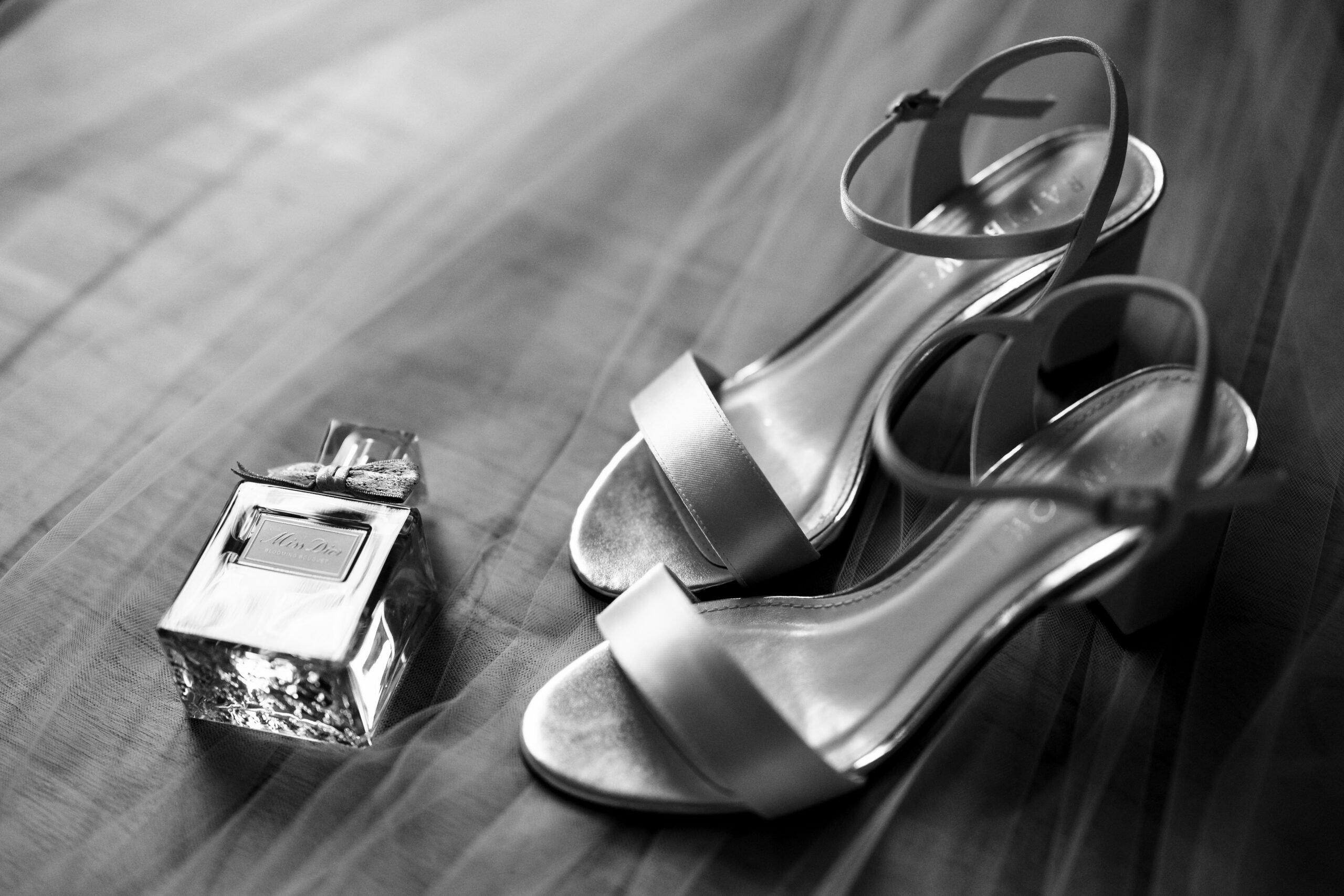 A black and white image of open toed, cream heeled shoes and a square bottle of Miss Dior perfume on top of a veil.