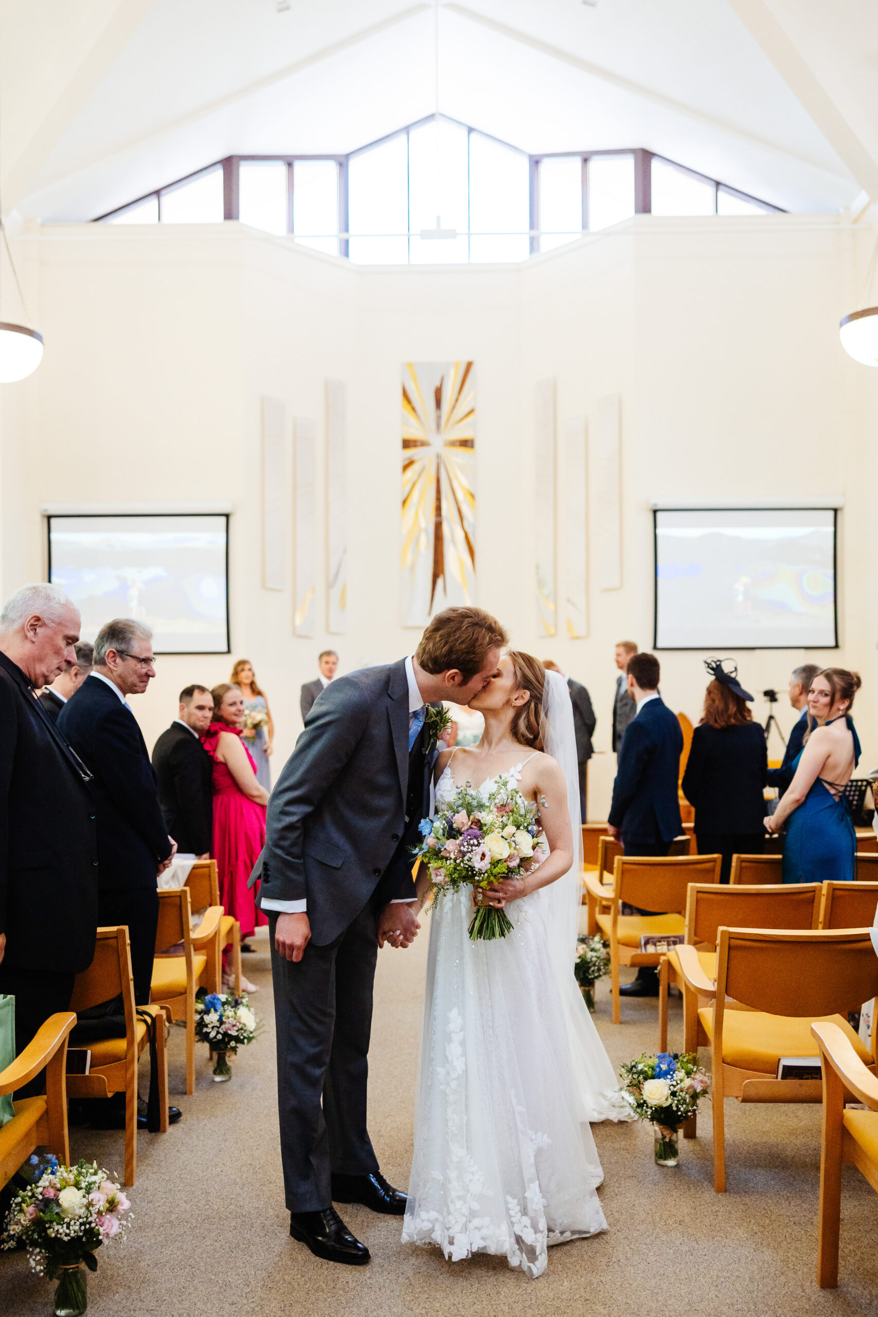 A photo of a bride and groom in the middle of the aisle kissing. The bride has her bouquet in her hands. They are in a church.