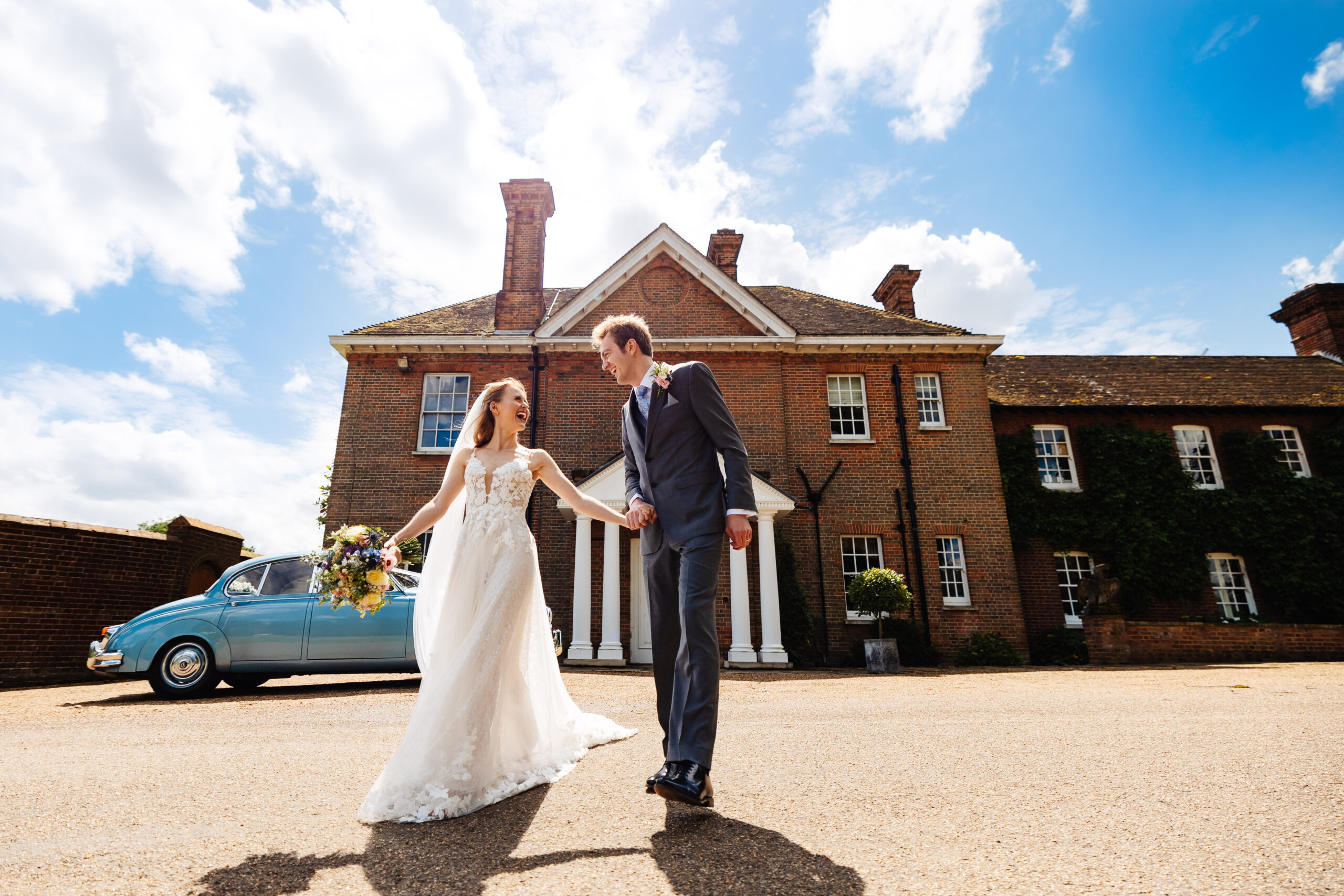 The bride and groom in front of a tall, old building in their wedding attire. They are holding hands and smiling at each other. The sky is bright blue with white clouds and you can see the blue wedding car in the background.