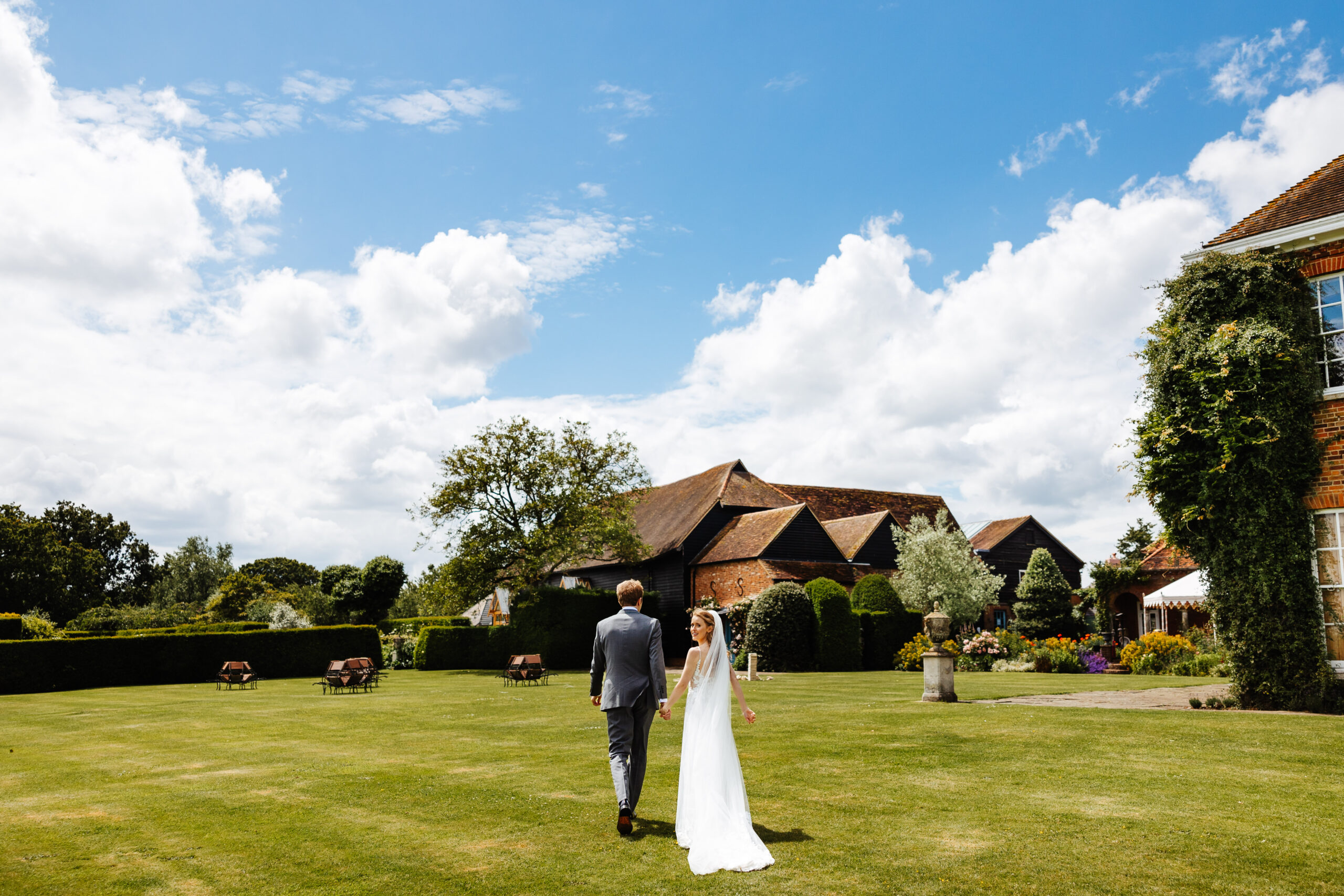 An image of an open garden and you can see the back of the bride and groom. They are holding hands and the bride is ever so slightly looking back over her shoulder and smiling at the camera.