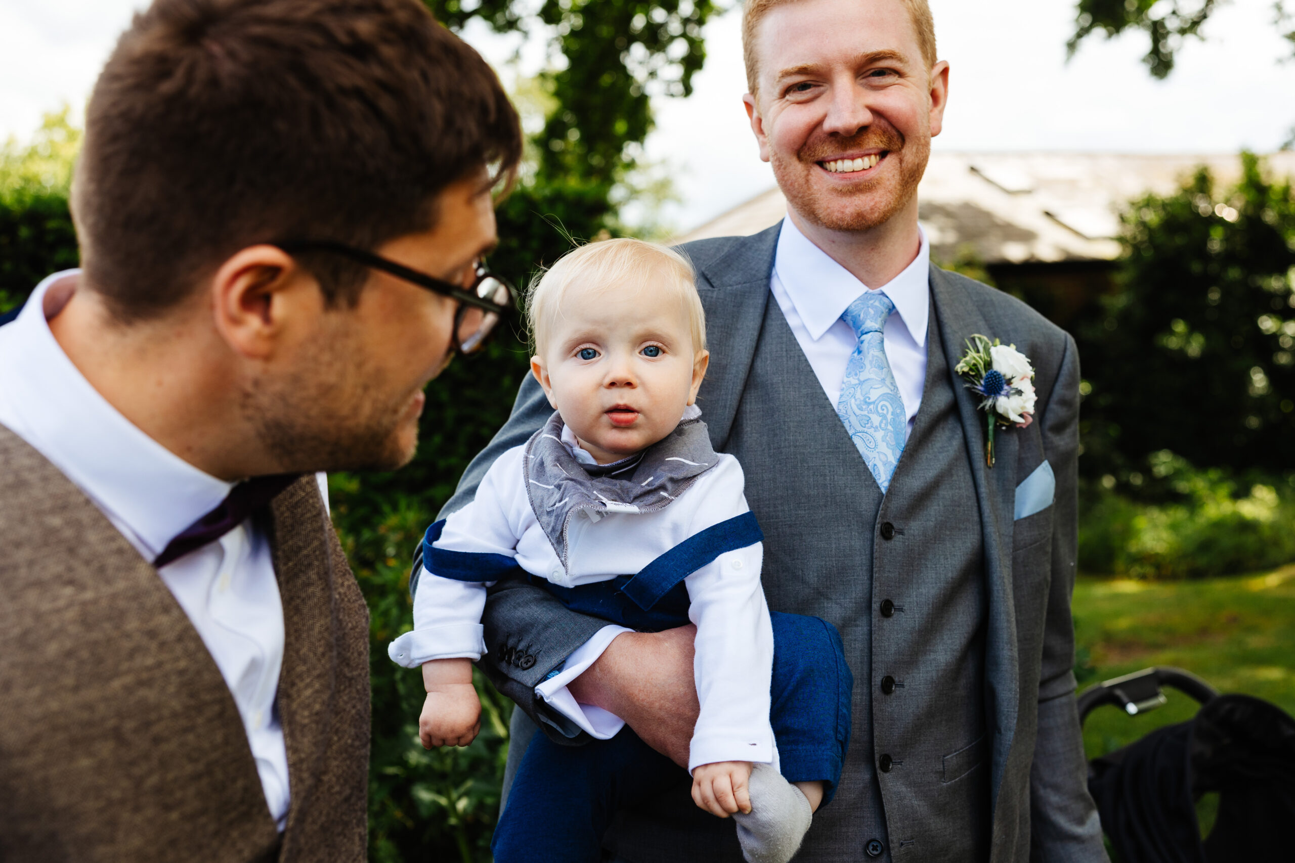 A man in a grey suit with blue detailing (tie and pocket square) holding a baby who is in a white shirt with blue braces. The man is smiling and the baby is looking at the camera with their bright blue eyes.