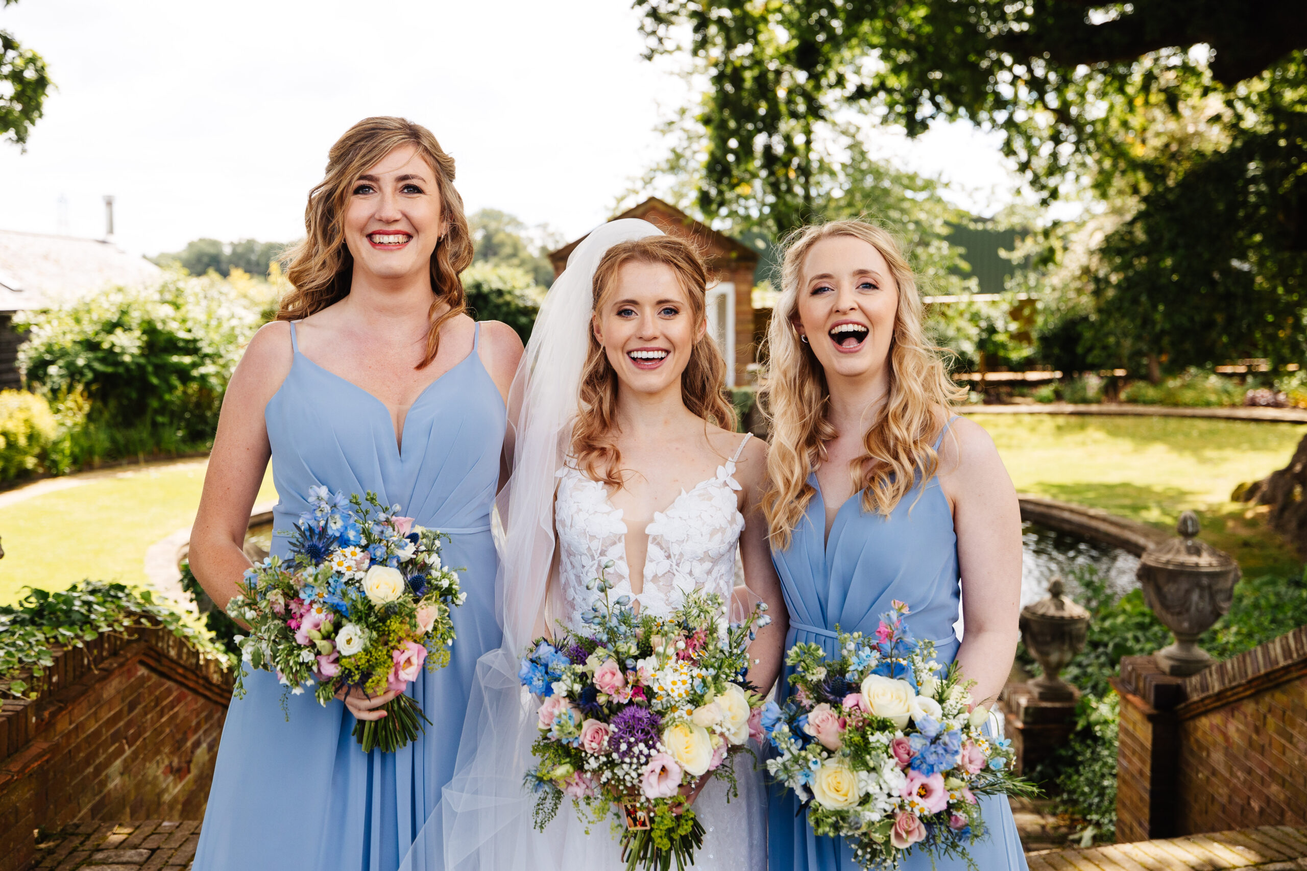 An image of a bride and her two bridesmaids standing next to her. They are all smiling and looking at the camera and holding their beautiful bouquets. The bridesmaids are in pale blue dresses.