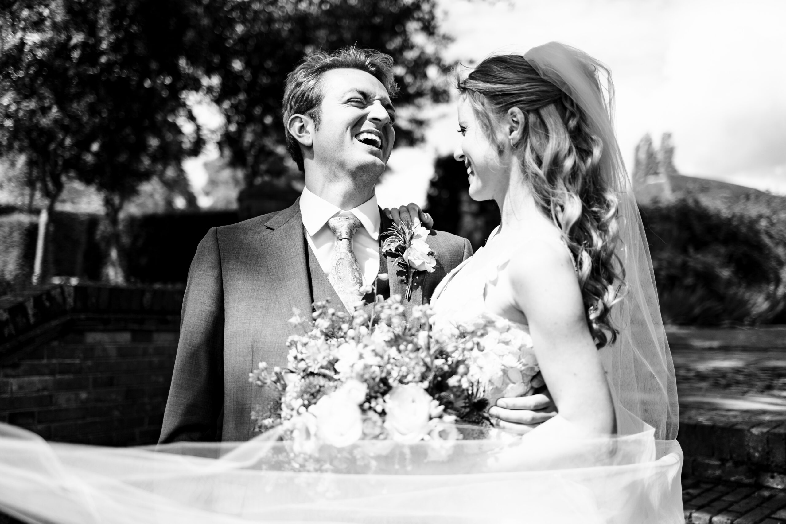 A black and white image of a bride and groom. The groom has his nose scrunched up laughing and the bride is looking at him smiling. Her veil is wrapped around her, blowing in the wind.