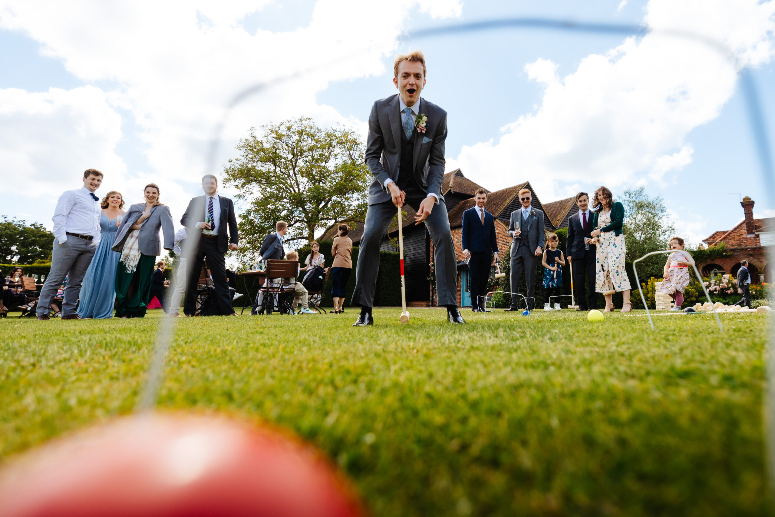 An image of the groom playing croquet. You can just about see the goal in the image as it's blurred out, but the groom is in the middle of it. He is holding a croquet stick and you can see the red ball in the bottom left of the image, albeit blurred.