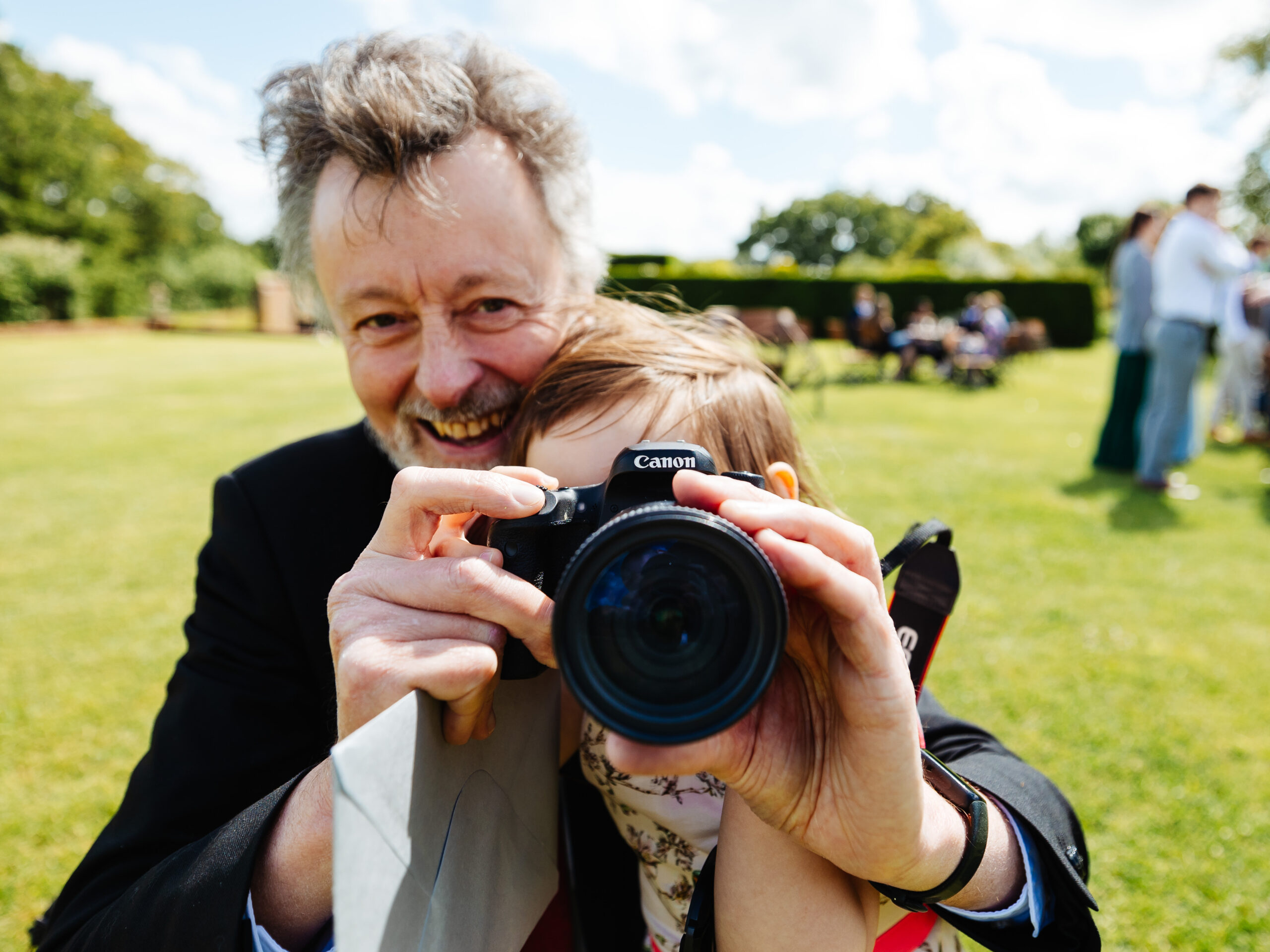 A picture of a boy in front of a man. The man is holding a camera in front of the boys face and is smiling. They are outside in the gardens.