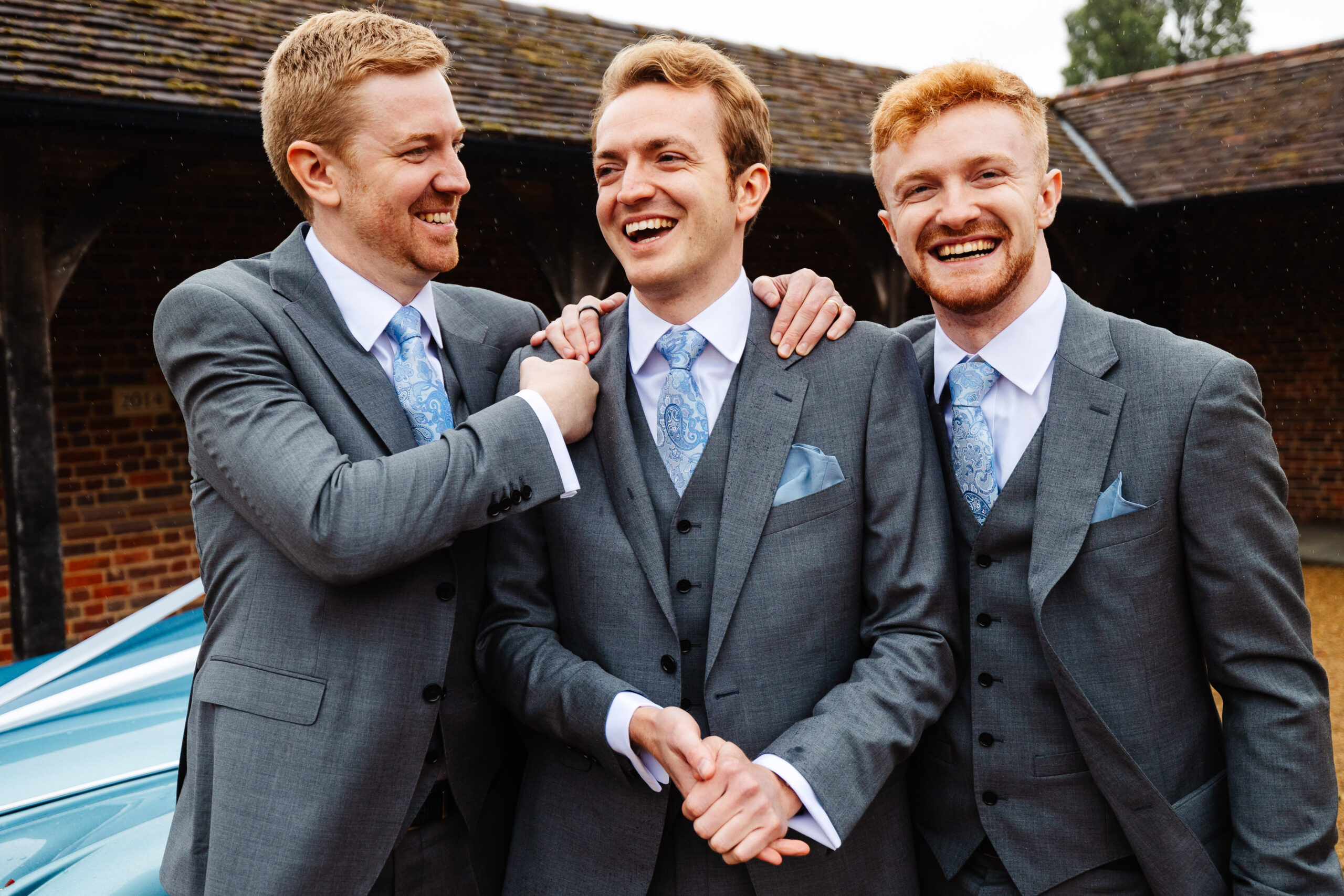 Three men in grey suits with blue details (ties, pocket squares). They are all smiling. One is looking at the camera, the other is looking at the man in the middle (the groom). The groom has his hands clasped together.