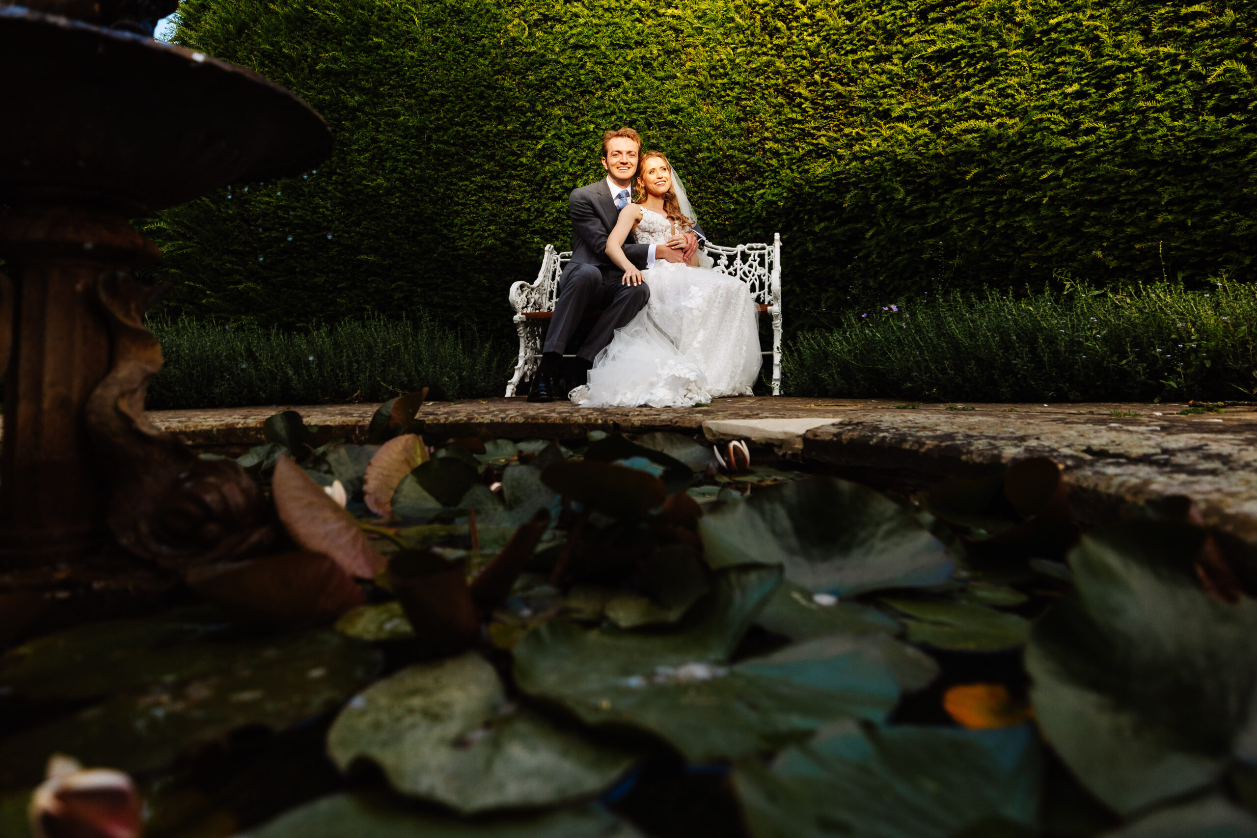 The bride and groom in front of a lily pond. They are sat on a bench and the bride has her hand on the groom's leg. They are looking at the camera and smiling.