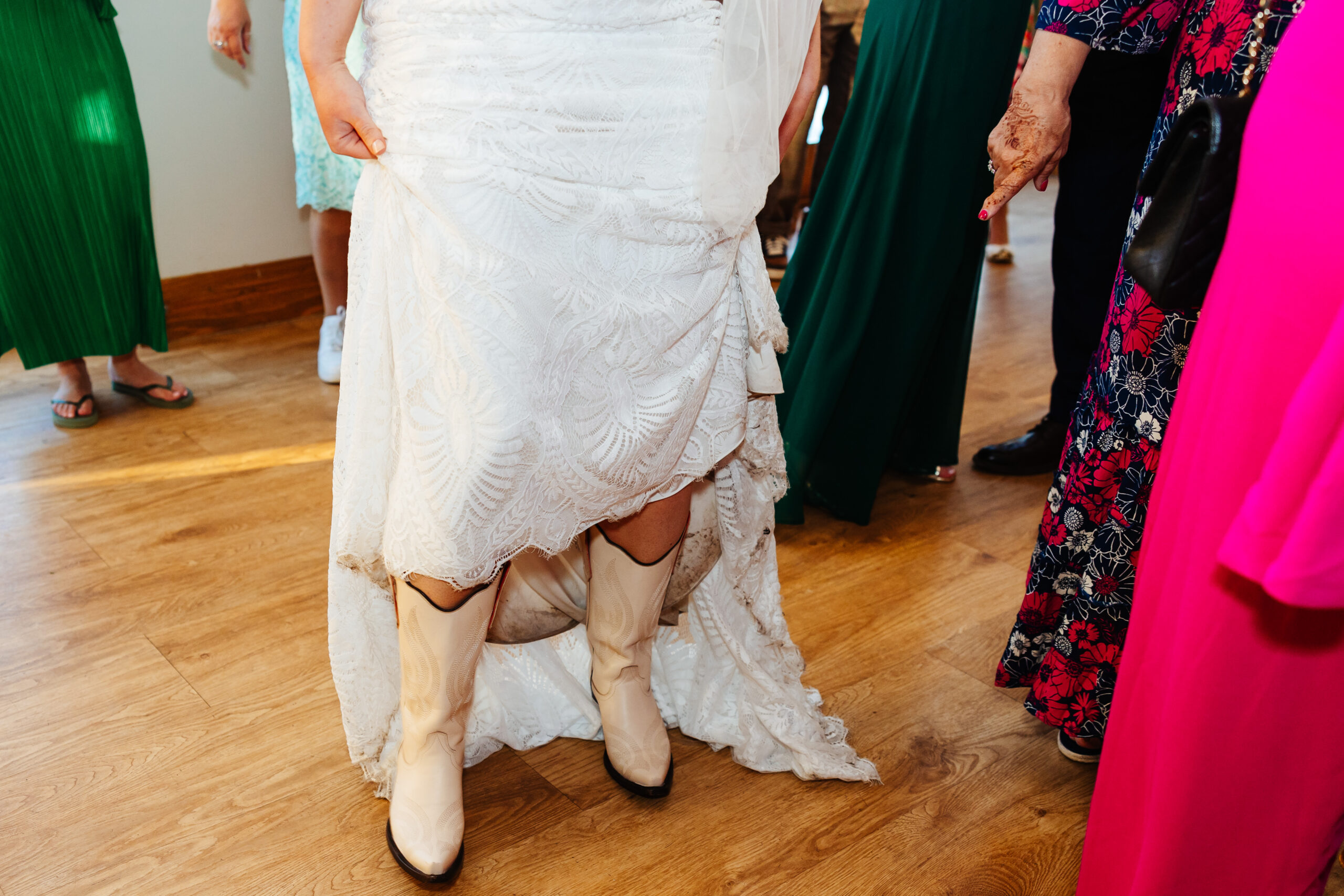 A photo of the bottom half of a bride in a wedding dress and she is wearing beige cowboy boots.