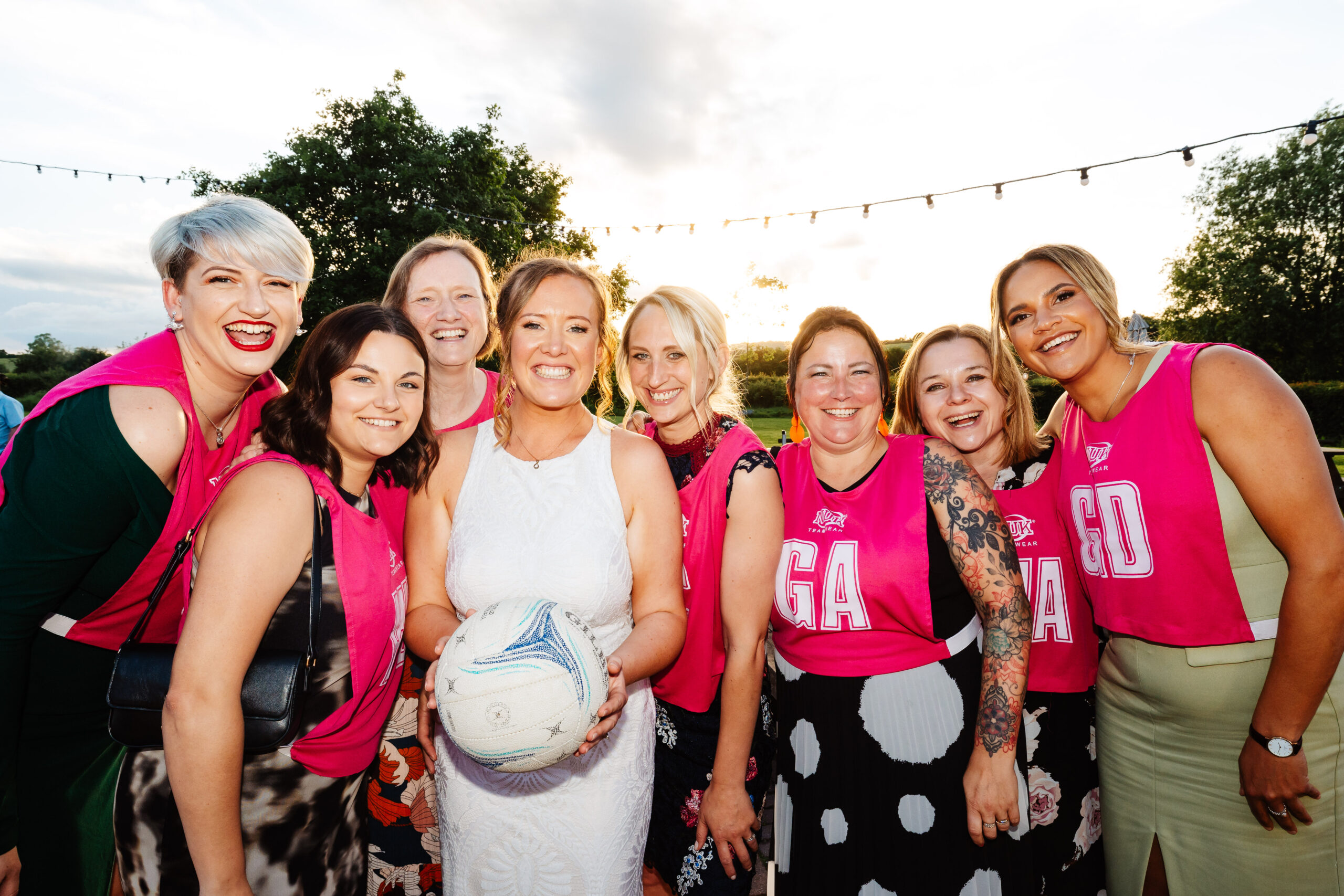 A group of ladies smiling with the bride in the middle. She is holding a netball and her friends are wearing bright pink netball vests.