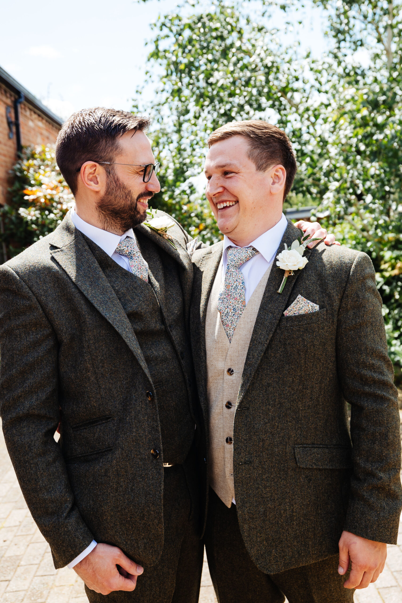 Two males looking at each other and smiling. One man has his arm around the other. They are both in green tweed suits.