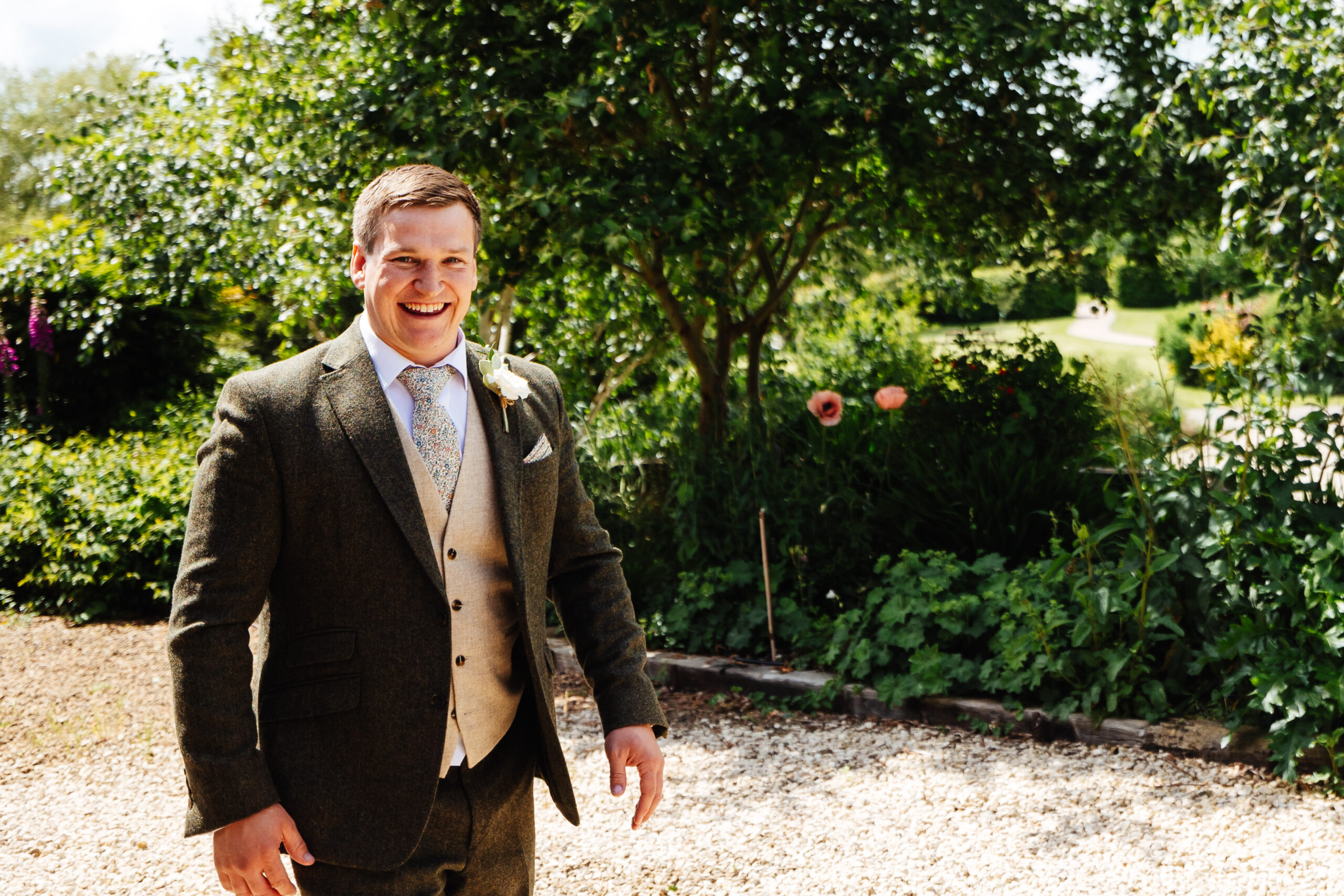 A groom wearing a green tweed suit with a beautiful landscape garden in the background. He is smiling and looks relaxed.