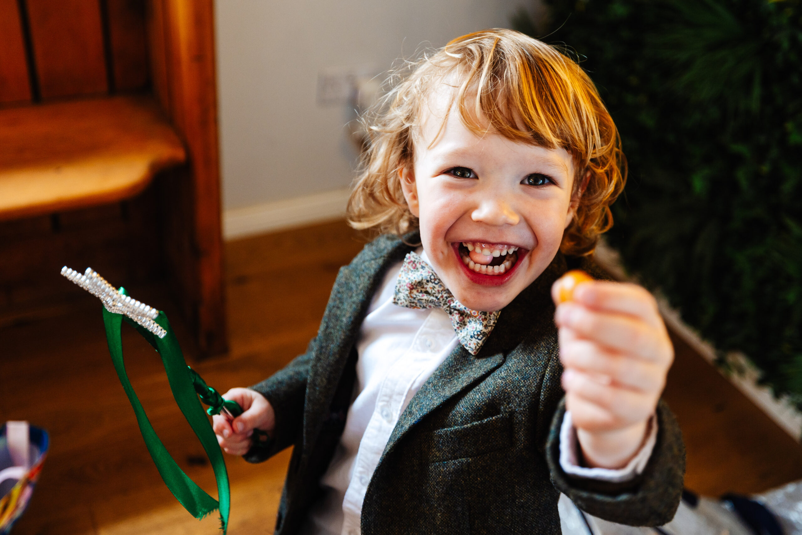 A little boy with a huge smile on his face. He is wearing a tweed jacket with a flower bow tie and has a sweet in his mouth and his hand. In the other hand he is holding a wand.