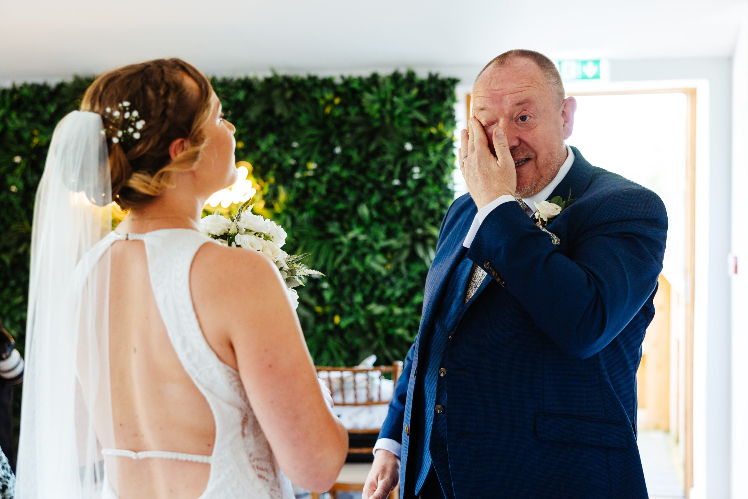 A bride and her father looking at each other. The father is drying his eyes after seeing his bride for the first time. The bride has her back to the camera and her dress is backless with a skinny white strap across the middle.