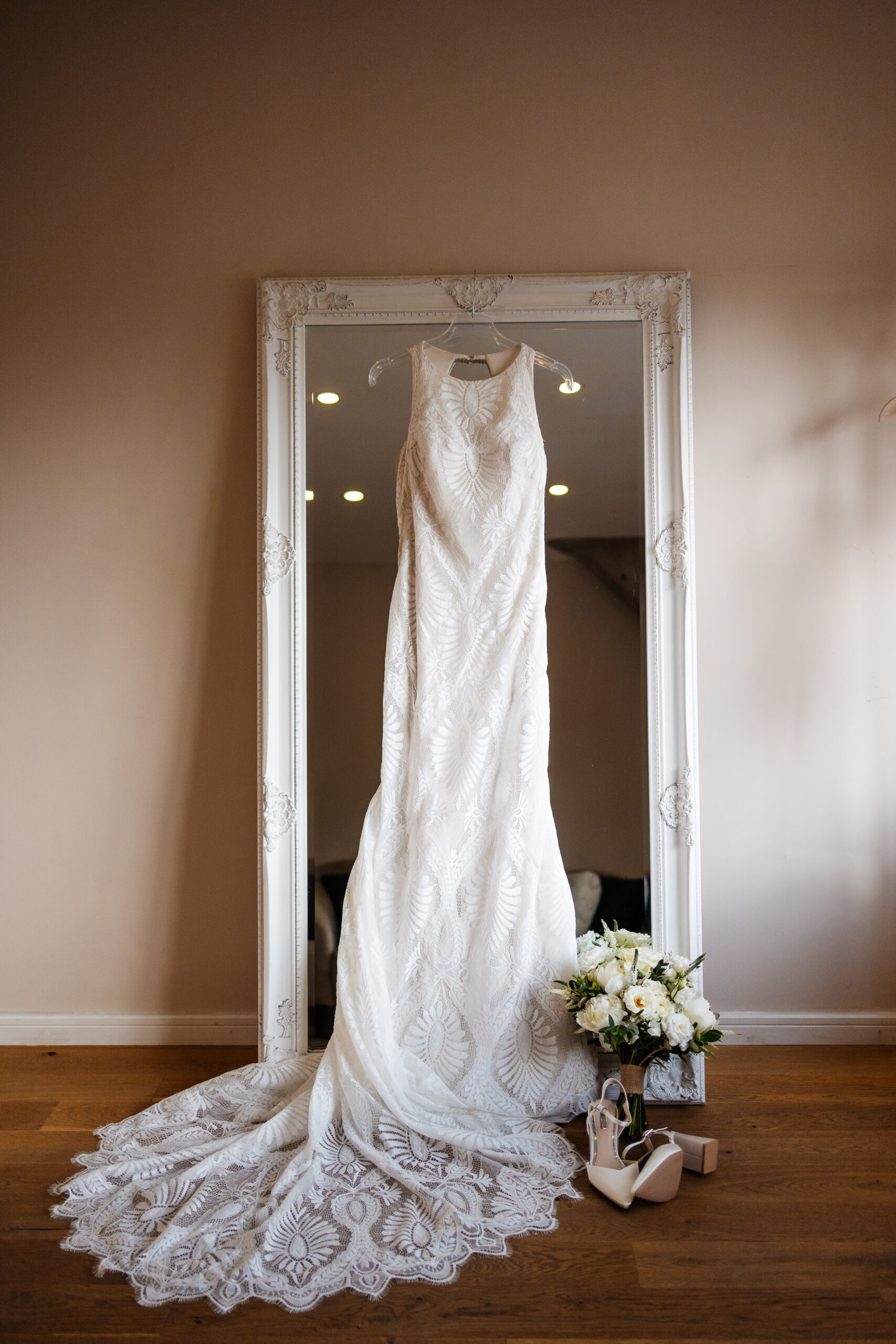 A photo of a long, white, lace wedding dress and white flowers in front of a long mirror with white edging. 