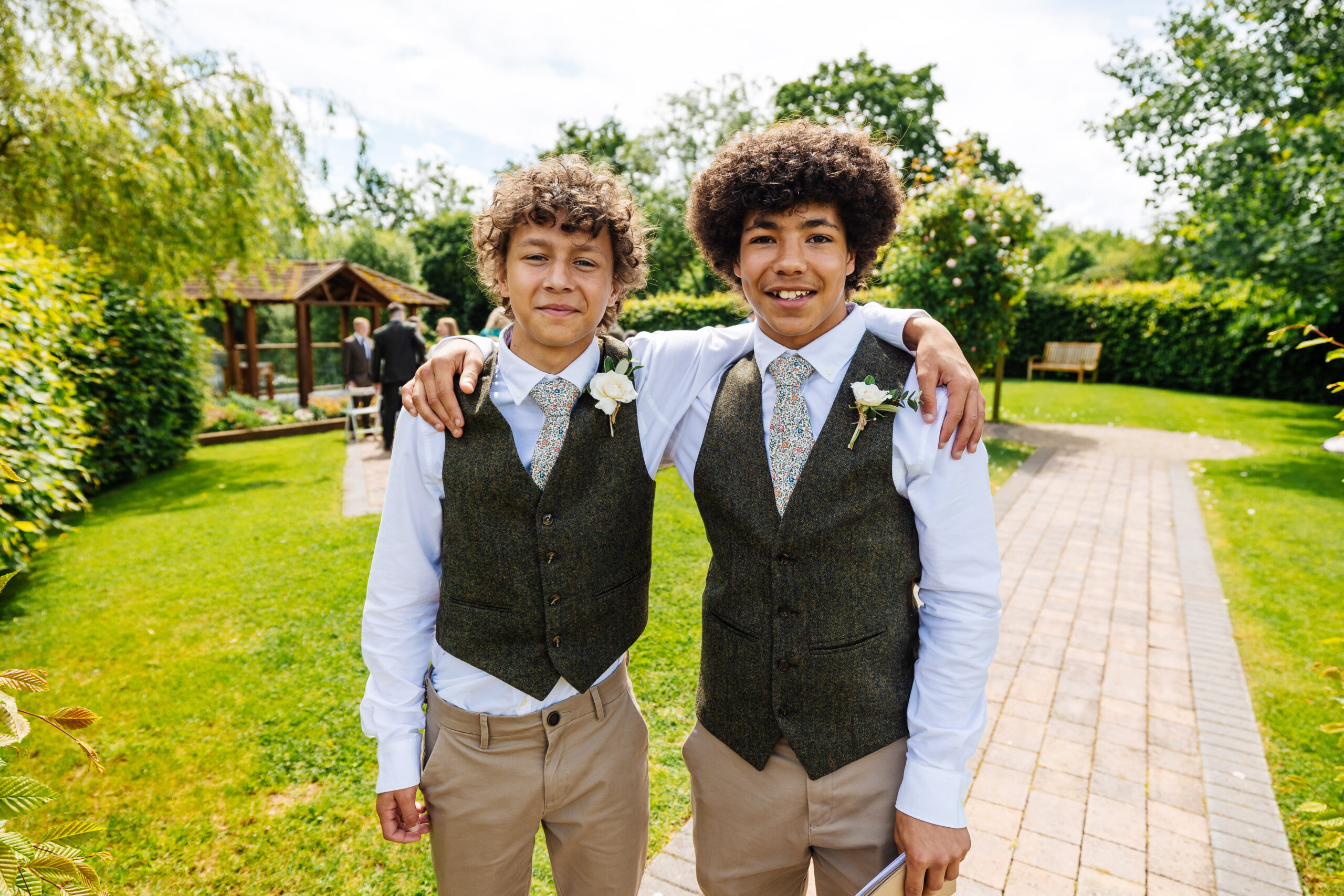 Two teenage boys wearing a tweed green waistcoats, white shirts, beige chinos and flowery ties. They are looking at the camera and smiling.