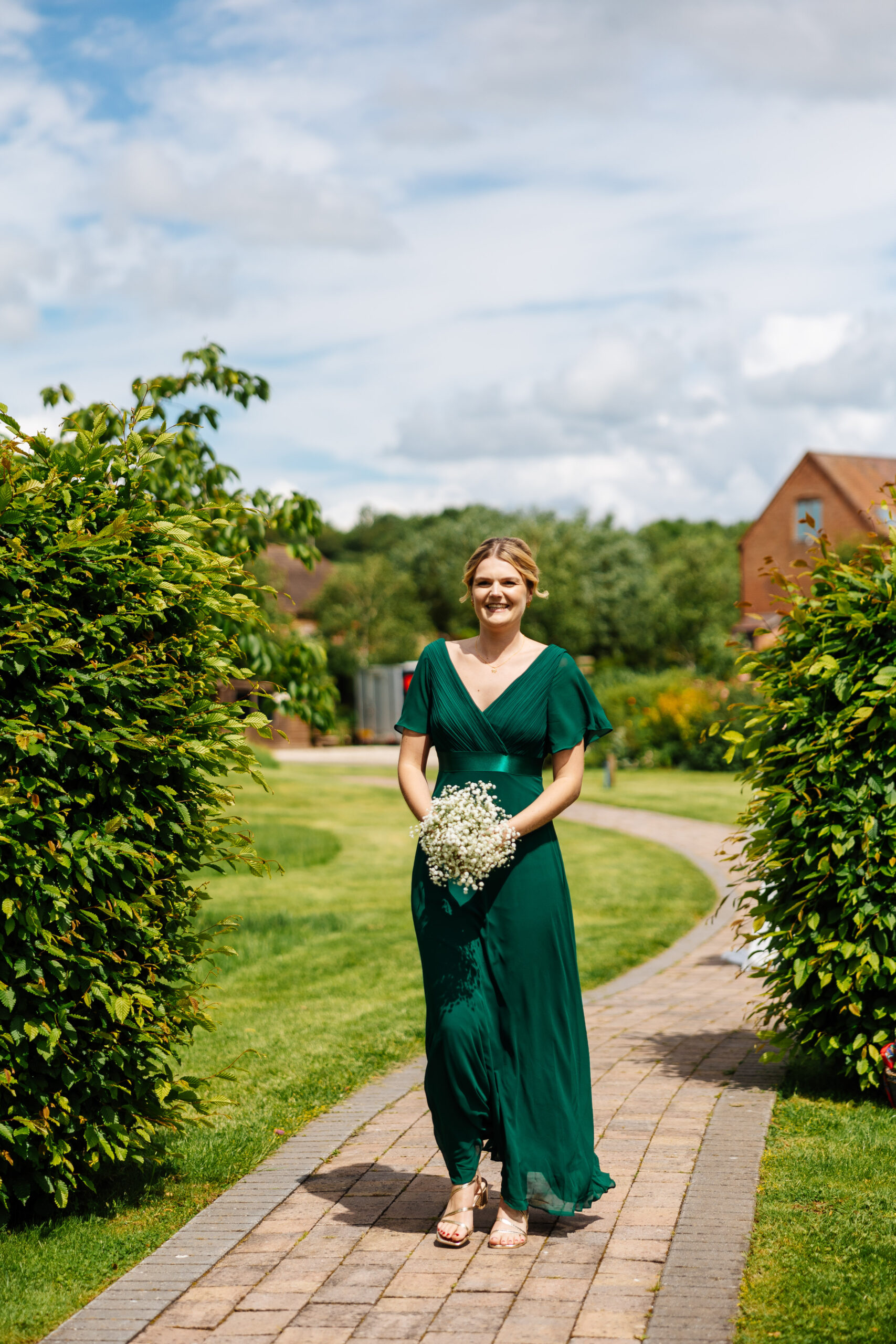 A blonde lady wearing a green dress holding a bunch of white flowers walking down a pavement with bushes either side.