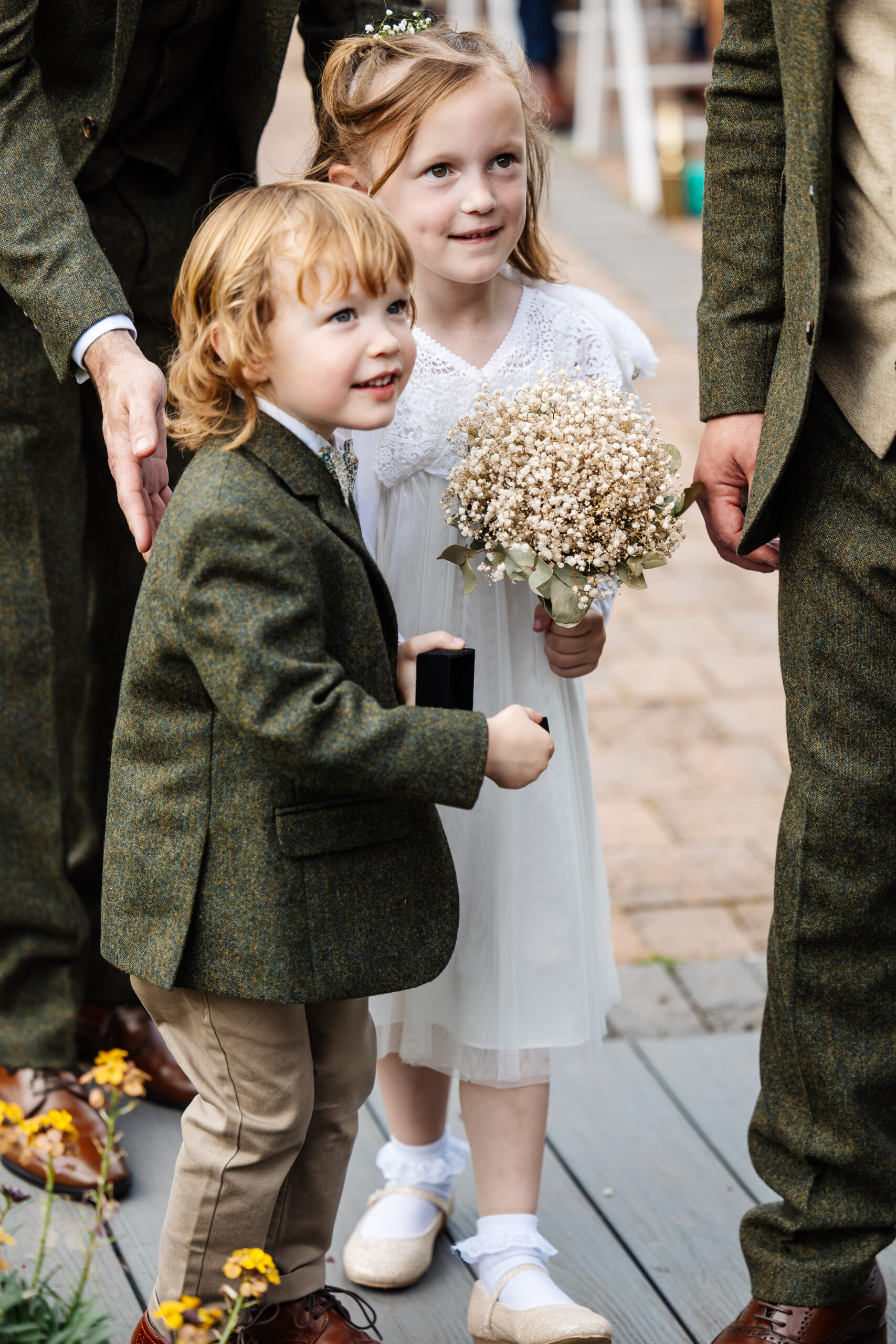 A little boy in a green tweed suit and a little girl in a white dress holding a bunch of flowers. They are looking away from the camera and are happy.