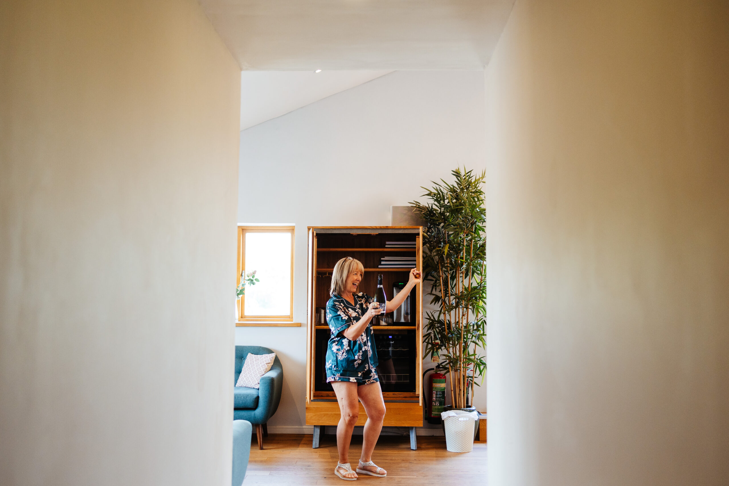 A photo of a lady in silk, blue pajamas holding a bottle of prosecco in one hand and her other arm in the air. She is happy.