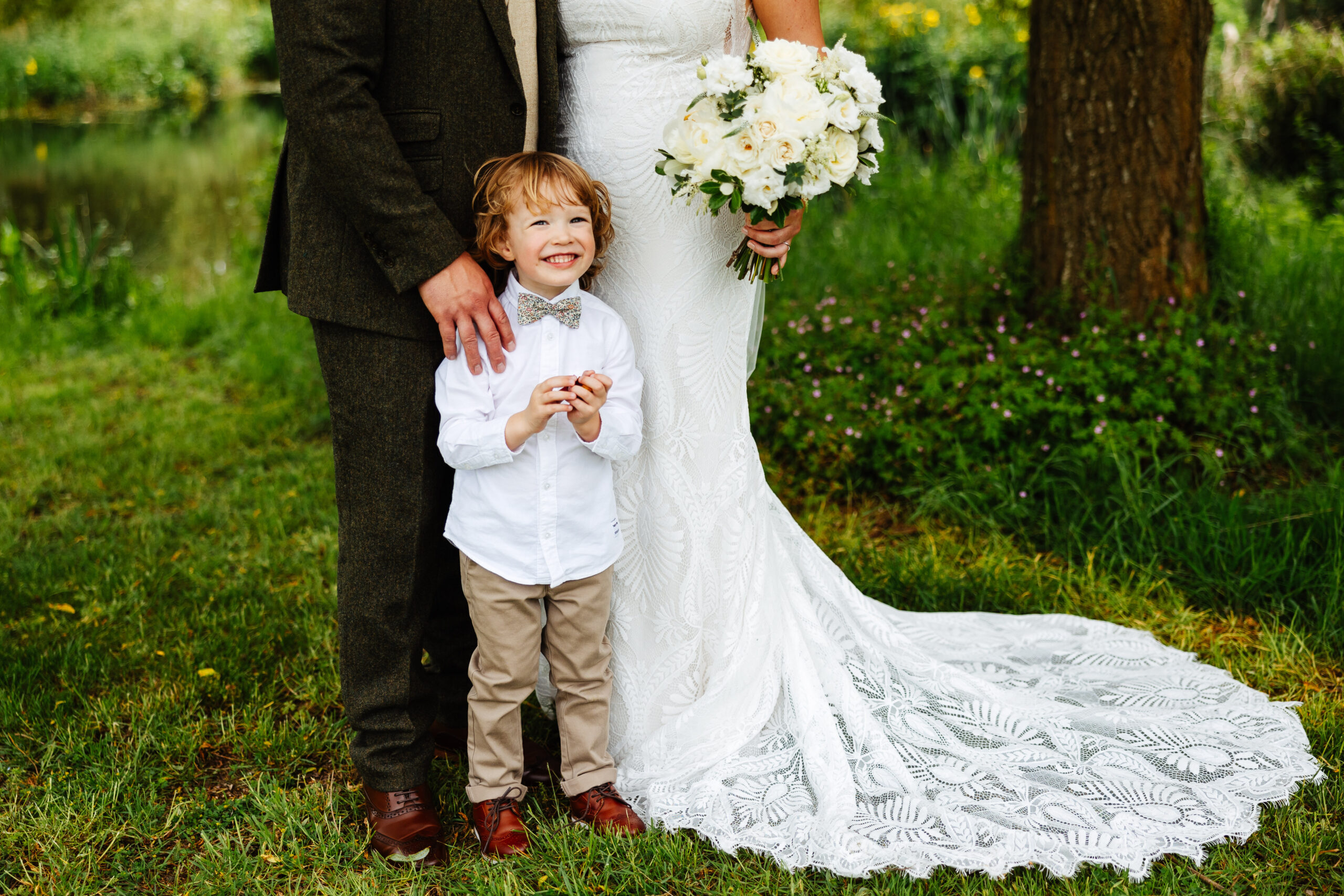 A photo of a little boy standing in front of his mum and dad who are the bride and groom. He is wearing a white shirt and beige chinos with a flowering bow tie and is smiling.