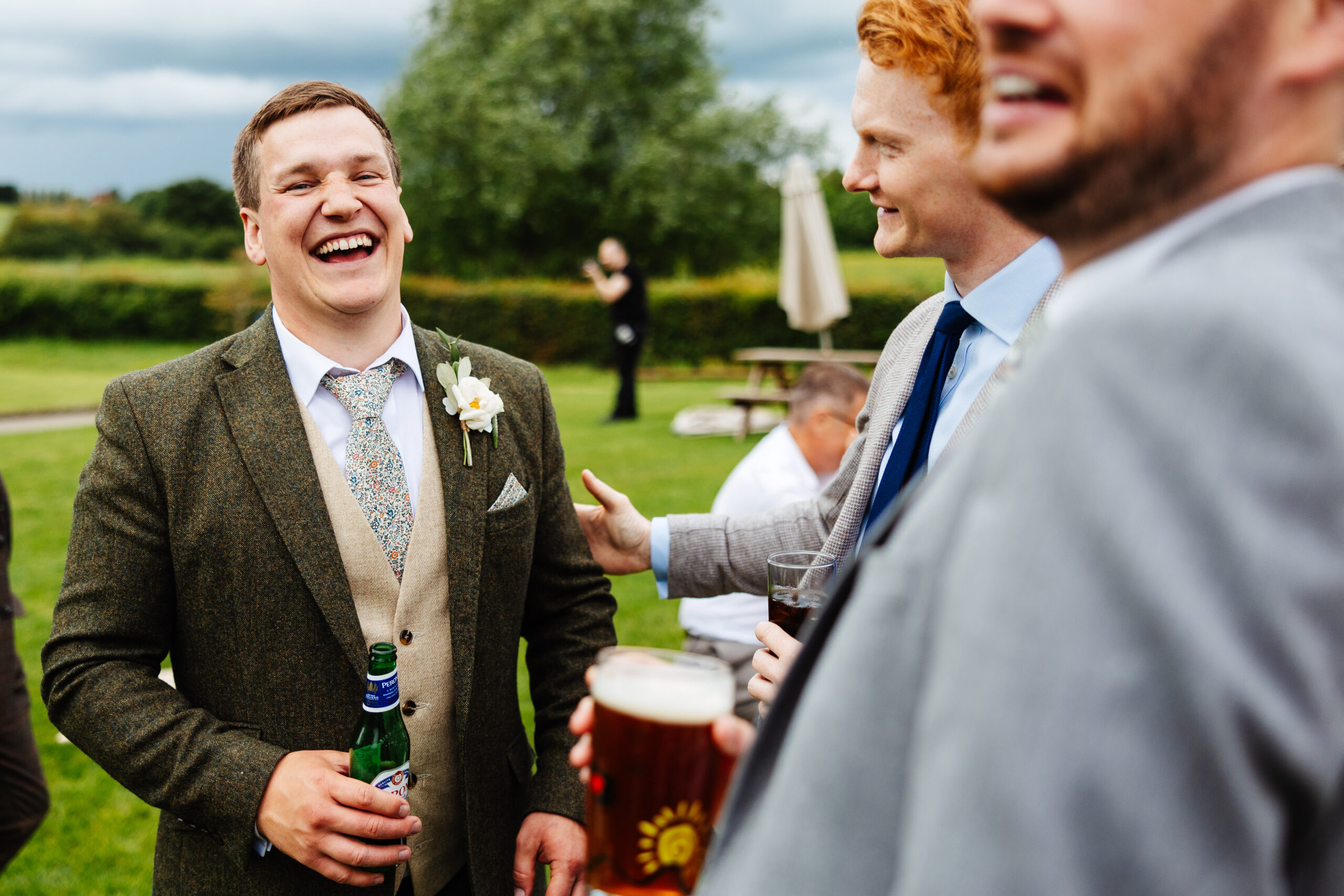 A groom in a tweed suit holding a beer and laughing. A man with red hair is smiling at him and holding his arm.