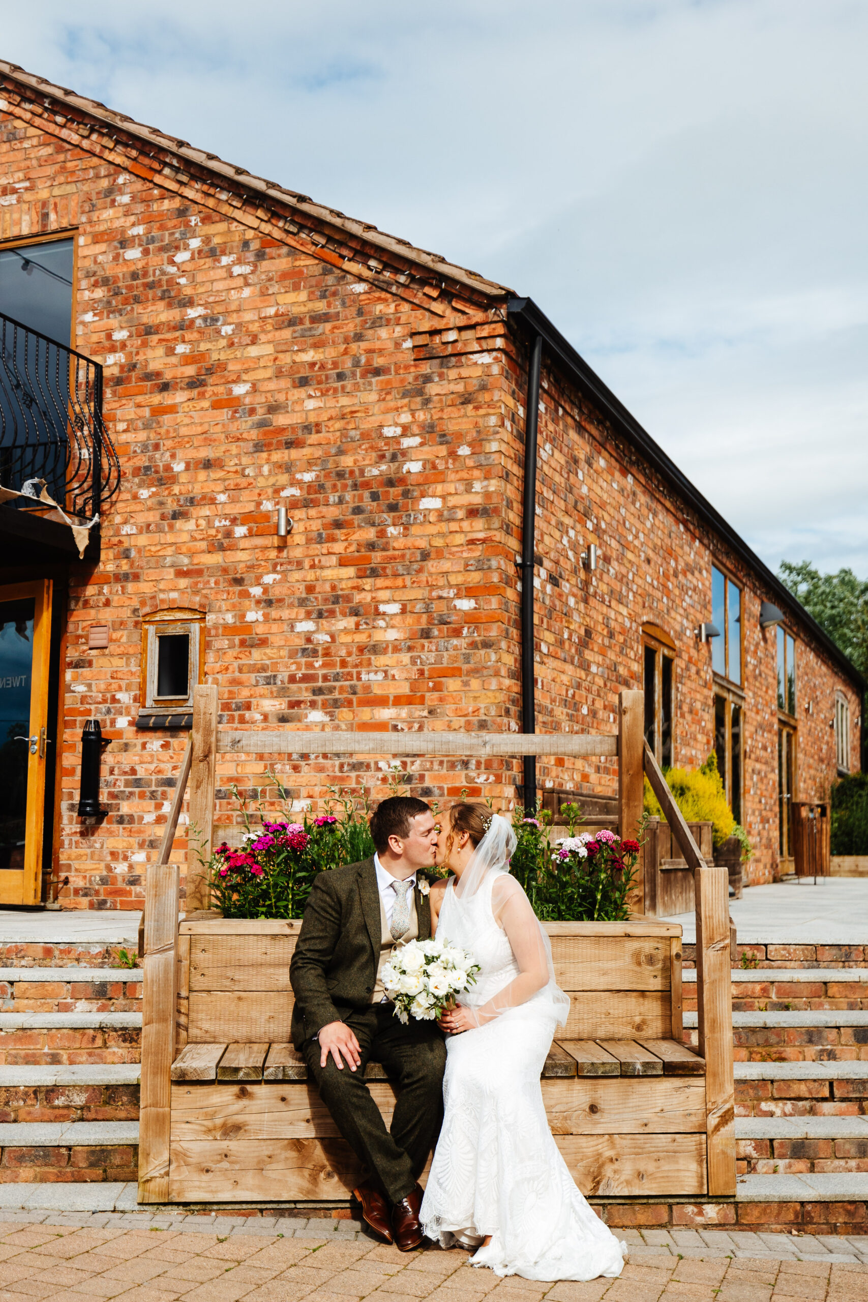 A bride and groom sat on a wooden bench infront of a brick building. The bride is holding a bunch of white flowers and wearing a white dress and veil. The groom is wearing a tweet suit and they are kissing.