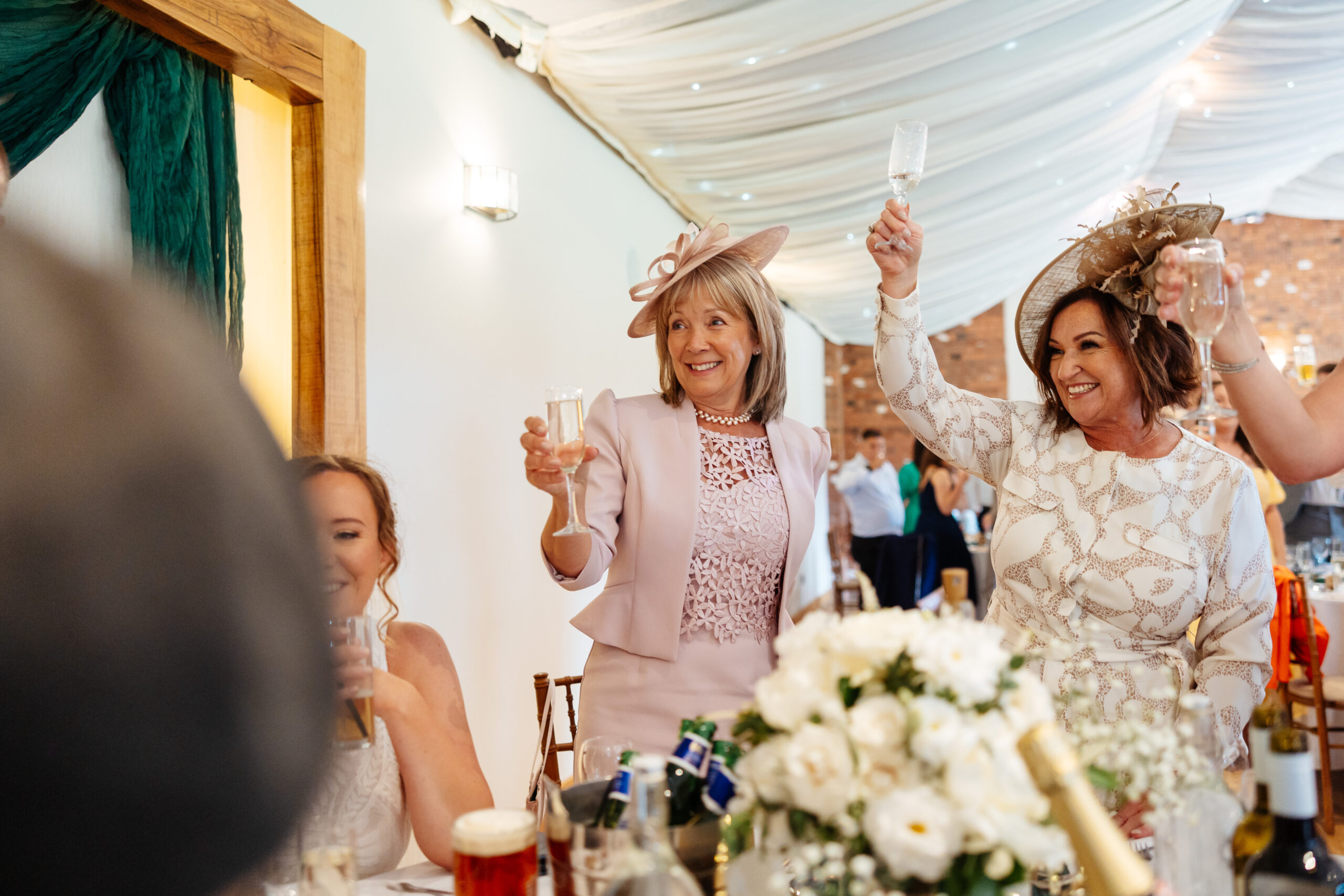 A photo of a two ladies; one in a pink suit with a hat and one with a white suit with a hat and they are holding a glass of prosecco up and smiling.