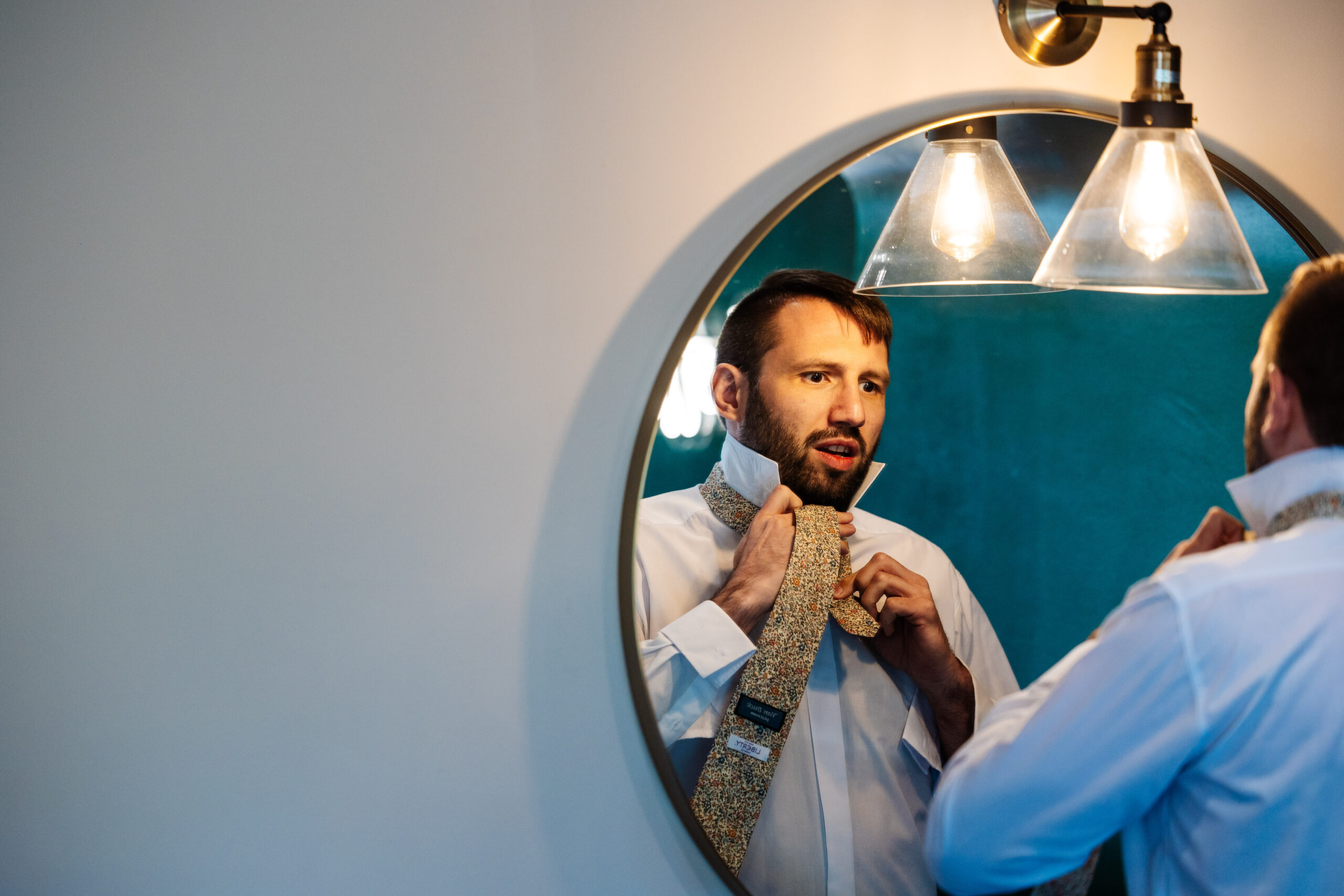 A photo of a man in a white shirt doing up his tie. He is looking in the mirror and you can see this in the image.