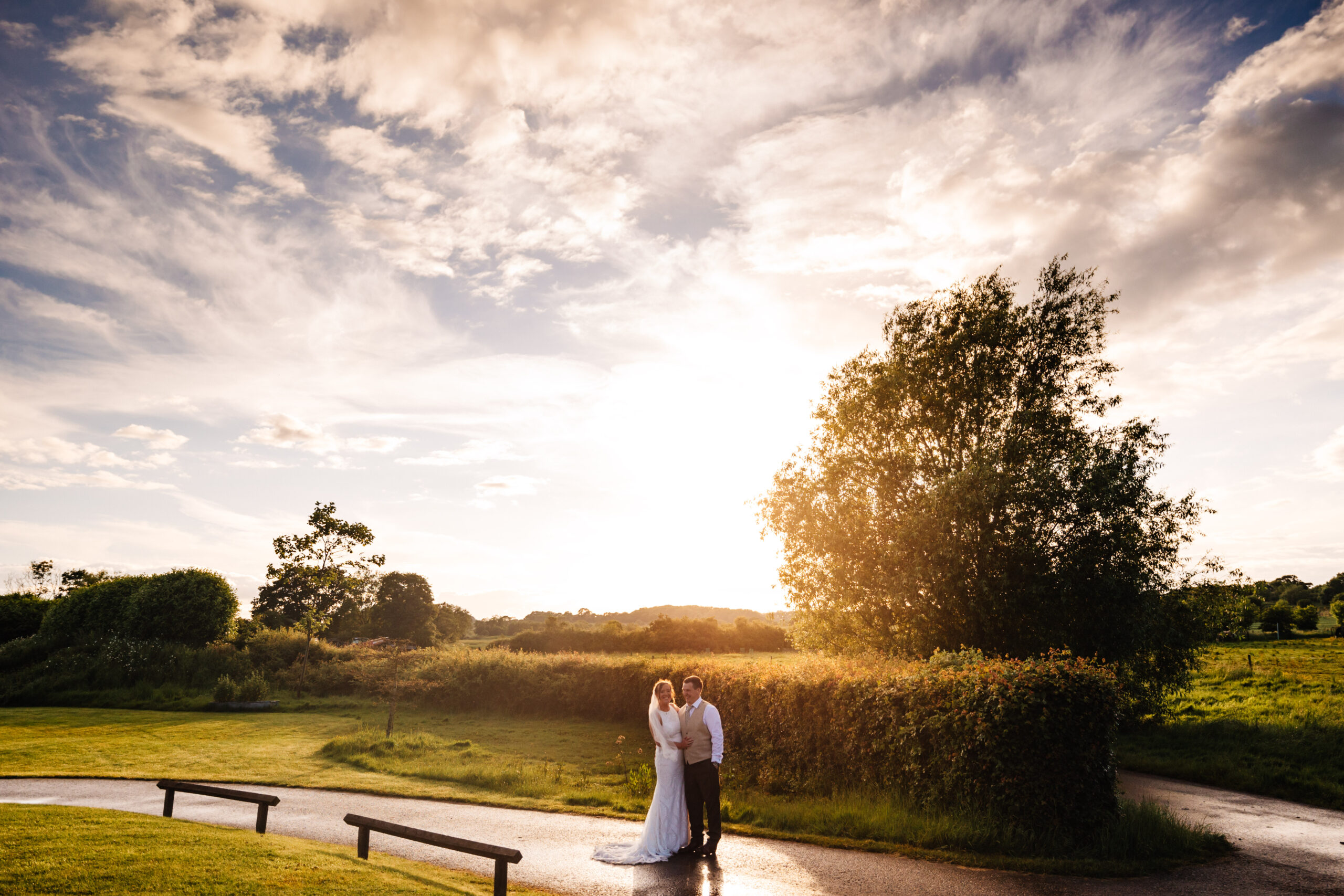 A bright blue sky with wispy white clouds and a field with trees and green grass. A bride and groom are standing in the middle smiling.